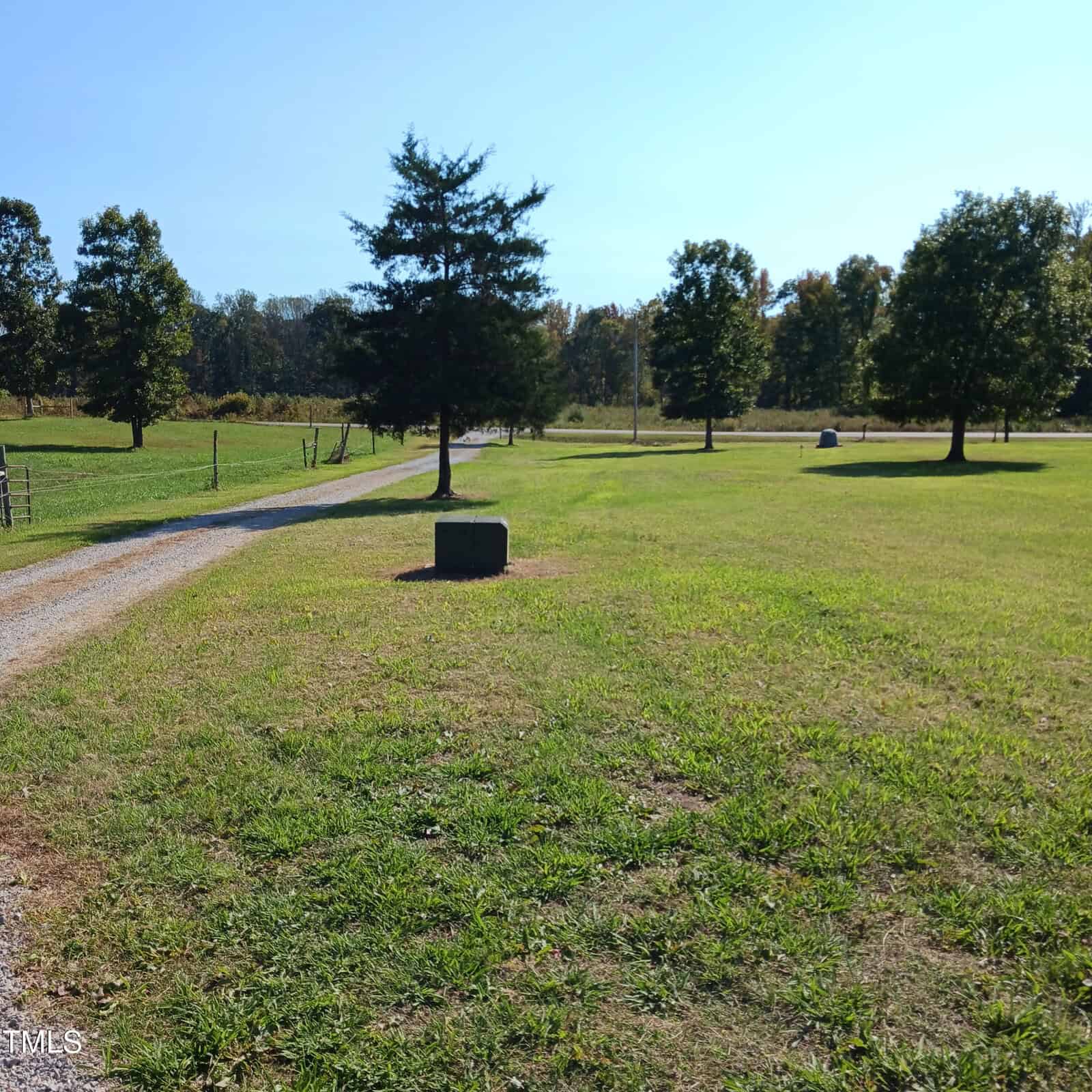 A grassy field with scattered trees and a gravel path leading into the distance. There is a concrete block near the center of the image, and a fence lines the left side of the path. The sky is clear and blue.