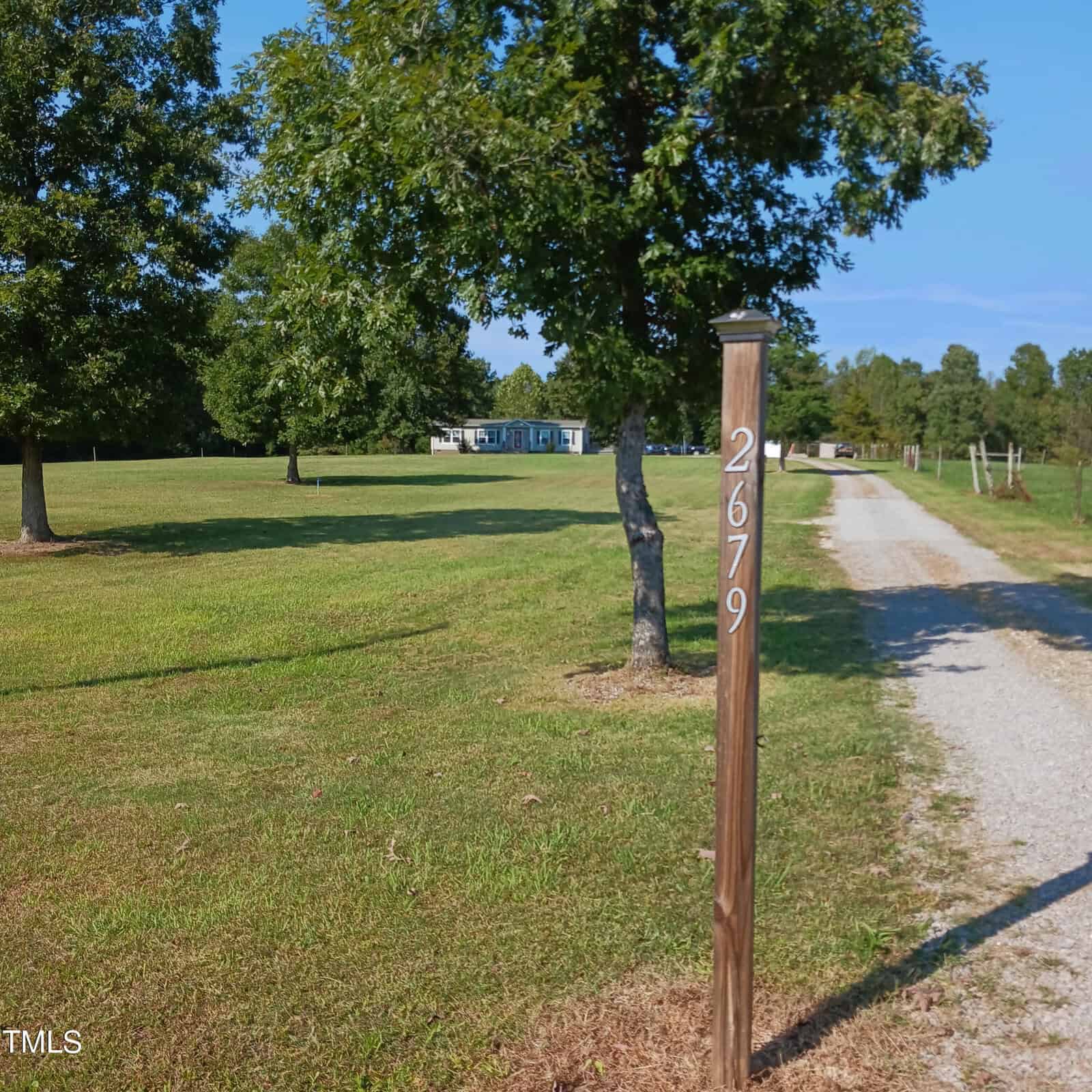 A gravel driveway leads to a house in the distance, bordered by a grassy lawn with trees. A wooden post beside the driveway displays the number 2679.