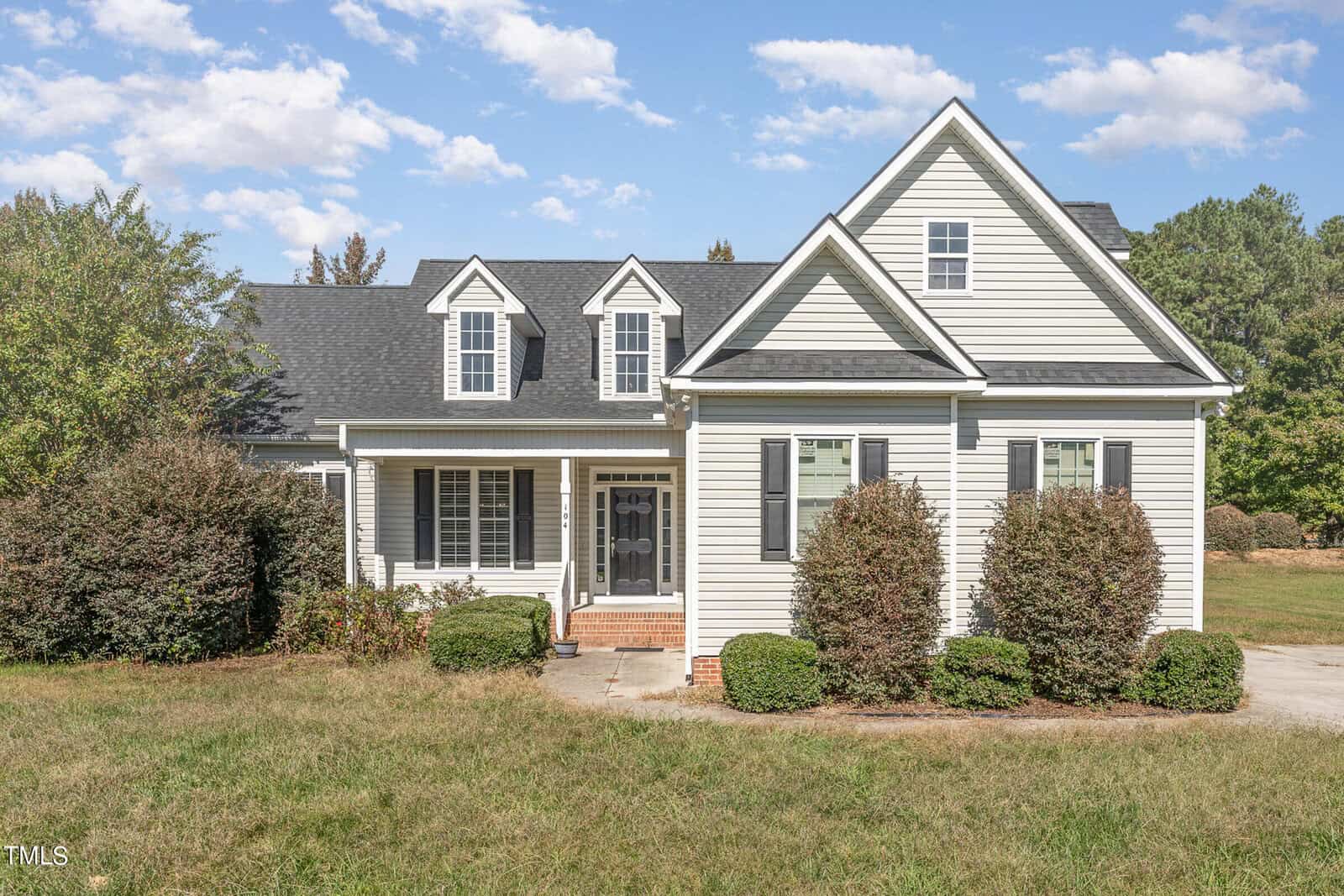 The white house at 104 Keeneland Drive boasts a gray roof, charming dormer windows, and a welcoming front porch with brick steps. Nestled in lush bushes and grass, this Oxford gem sits under a clear blue sky with scattered clouds.