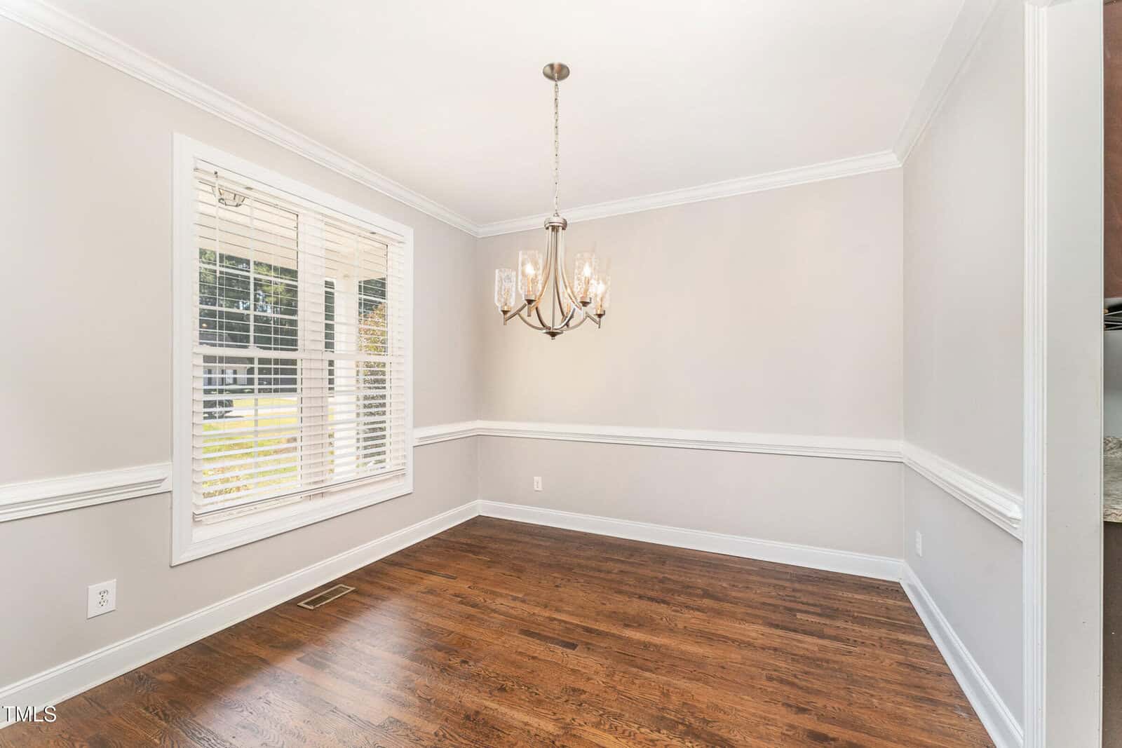 A bright, empty dining room with light gray walls, white trim, and hardwood floors. A chandelier hangs from the ceiling. Sunlight streams through a window with white blinds.