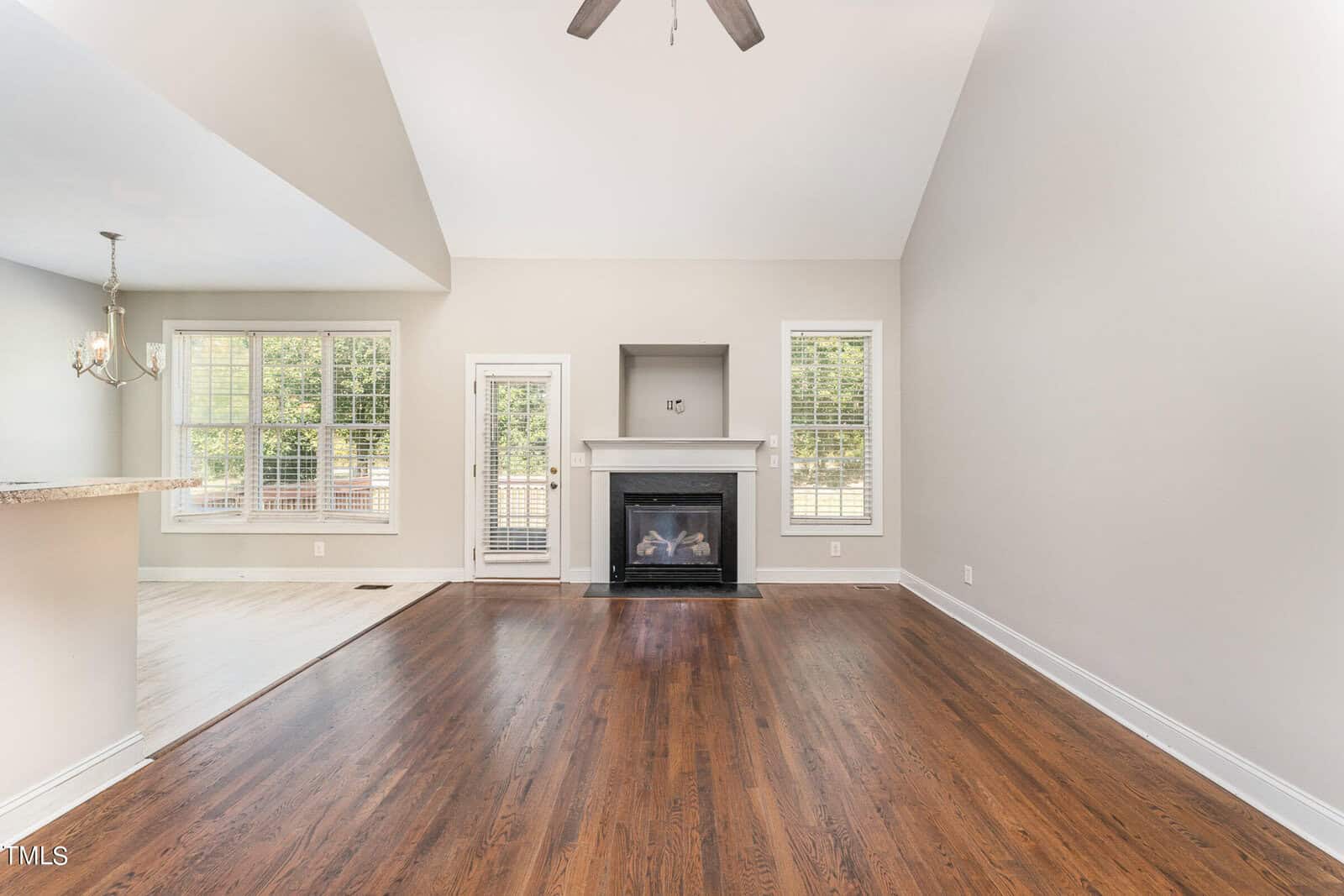 A spacious living room features a high ceiling, hardwood floors, and a central fireplace flanked by windows. Natural light fills the room from large windows and a glass door leading to an outdoor area.