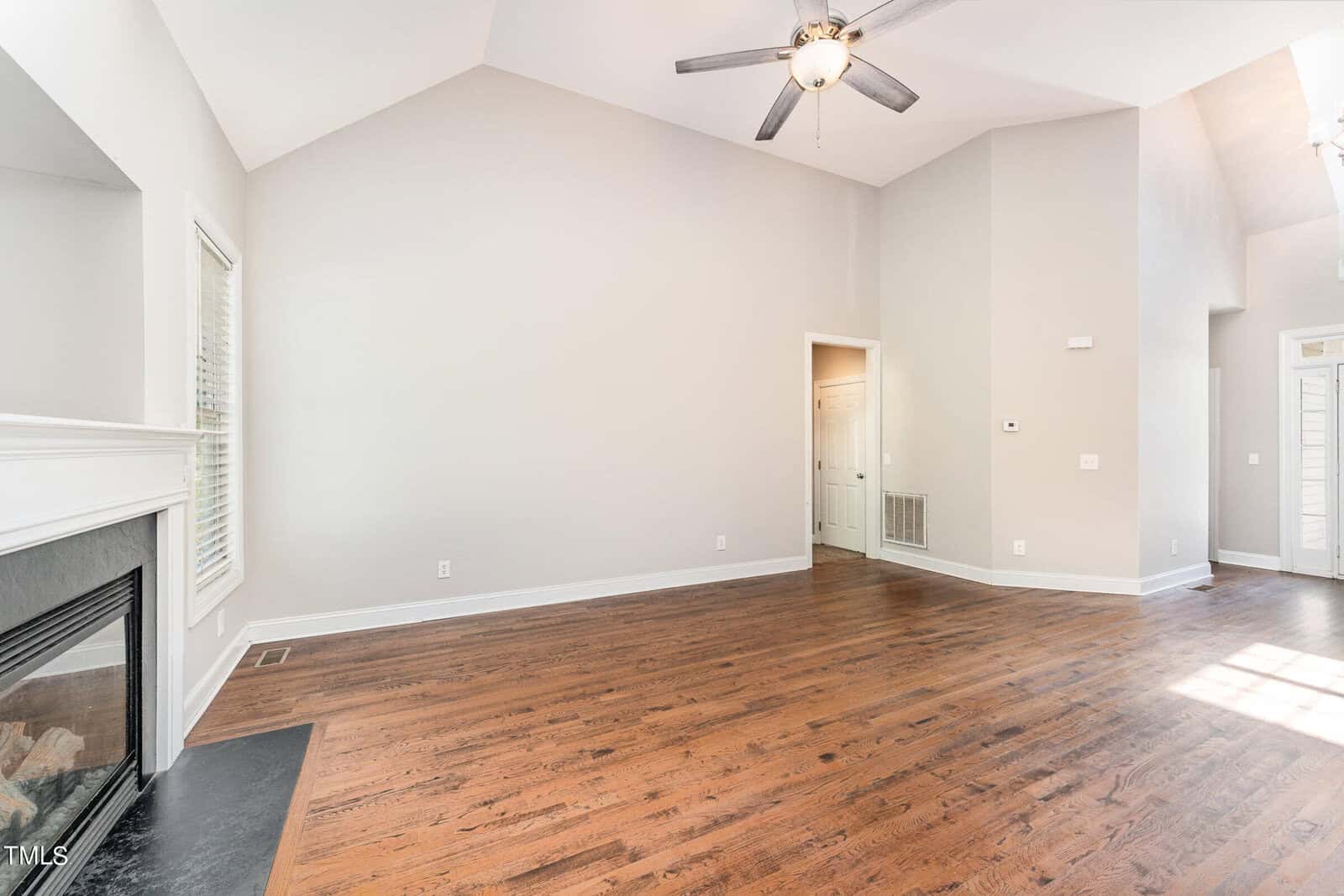 A spacious, empty living room with hardwood floors, a ceiling fan, and neutral-colored walls. A fireplace is in the corner, and a hallway leads to other rooms. Large windows allow natural light to fill the space.