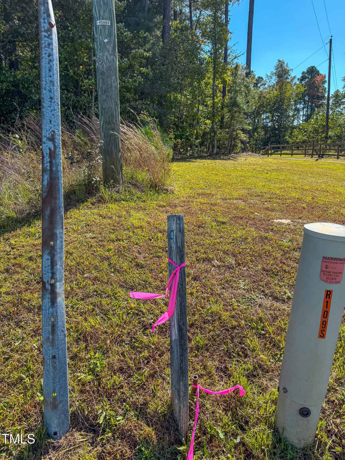 A grassy field with pink ribbon tied around a short wooden stake. Nearby, there's a tall metallic pole and a utility pole. In the background, trees line the field under a clear blue sky.