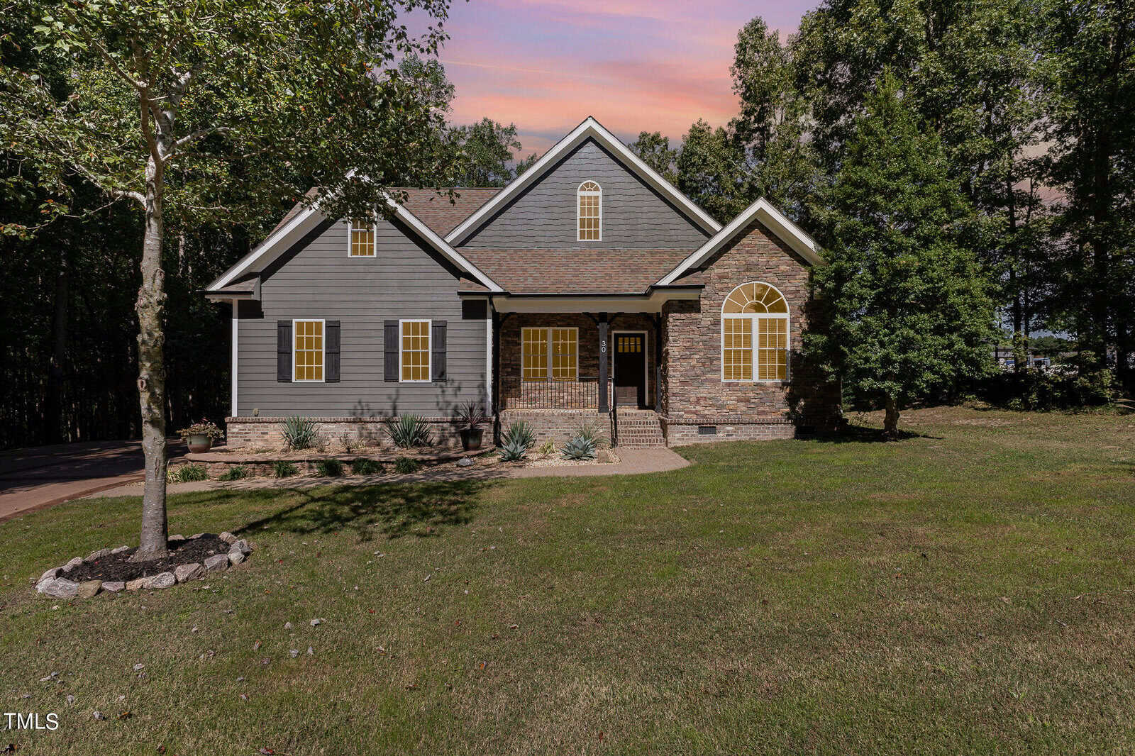 A single-story gray house with yellow-trimmed windows and a brick entryway, surrounded by trees and a large green lawn, under a colorful sunset sky.