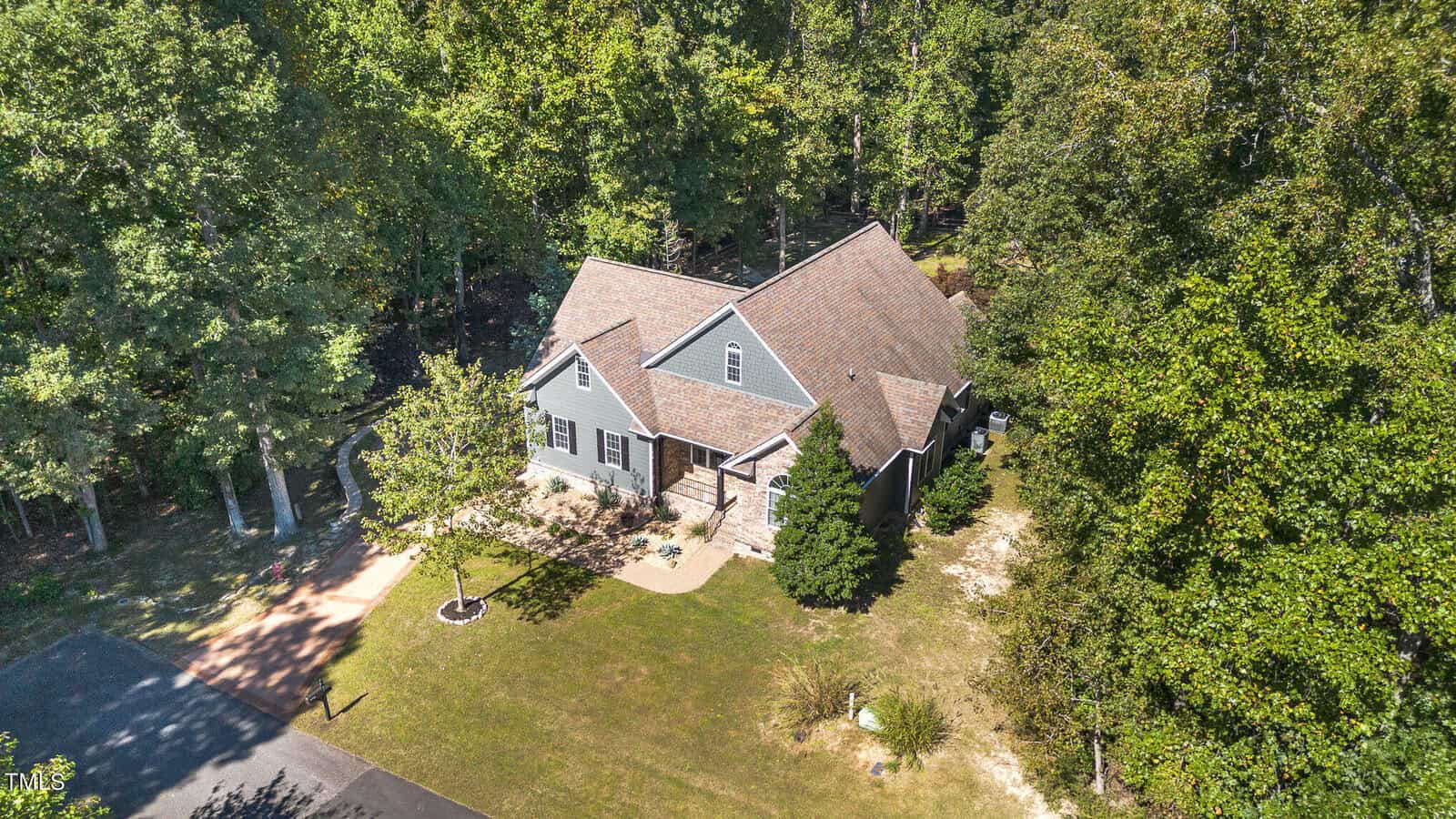 Aerial view of a light gray house with a brown roof, surrounded by trees and greenery. The house has a small front yard with a path leading to the entrance, and it sits next to a quiet road.