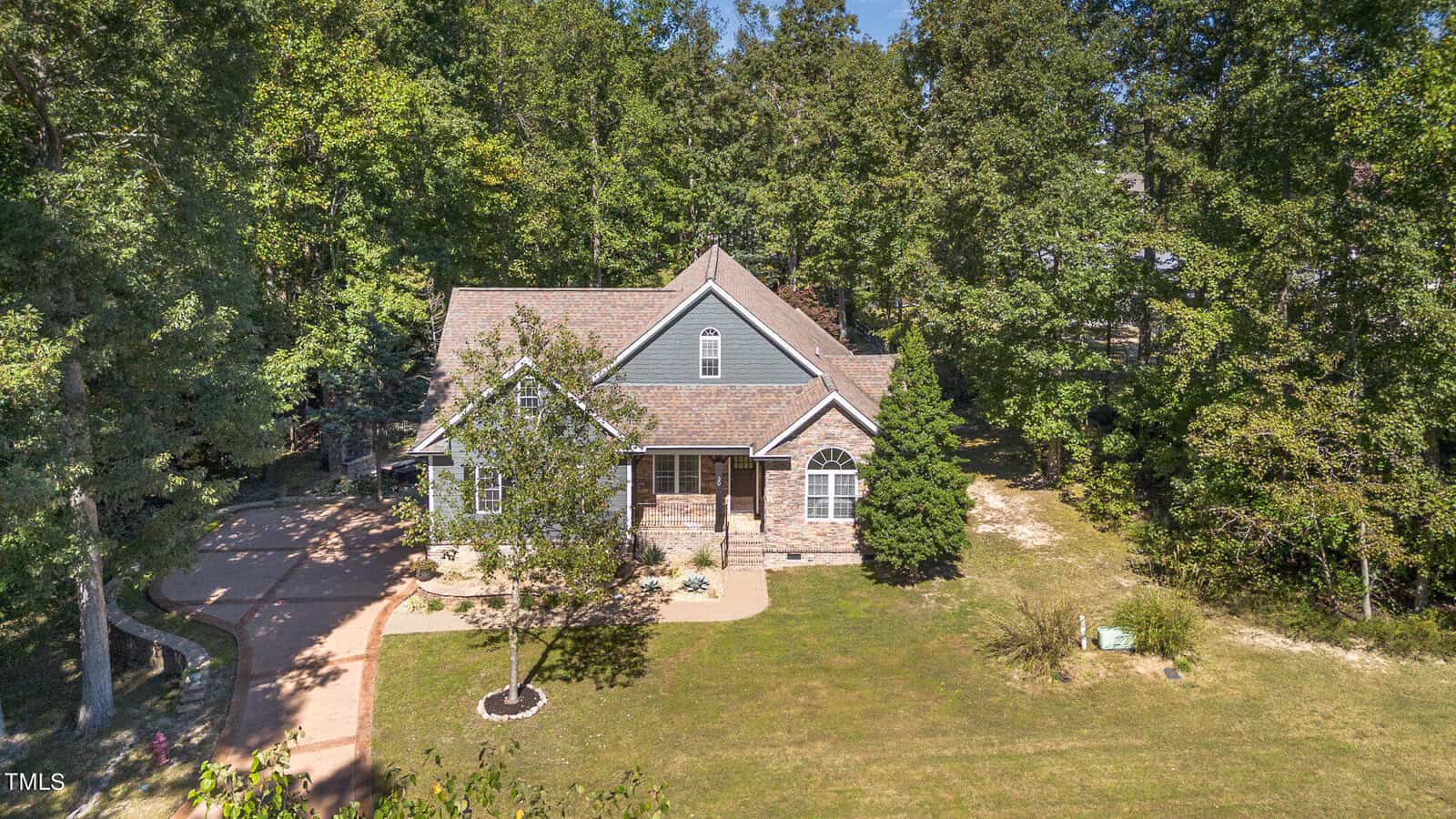 Aerial view of a two-story house with gray siding and stone accents, surrounded by trees and a spacious front yard with a curved driveway and landscaping.