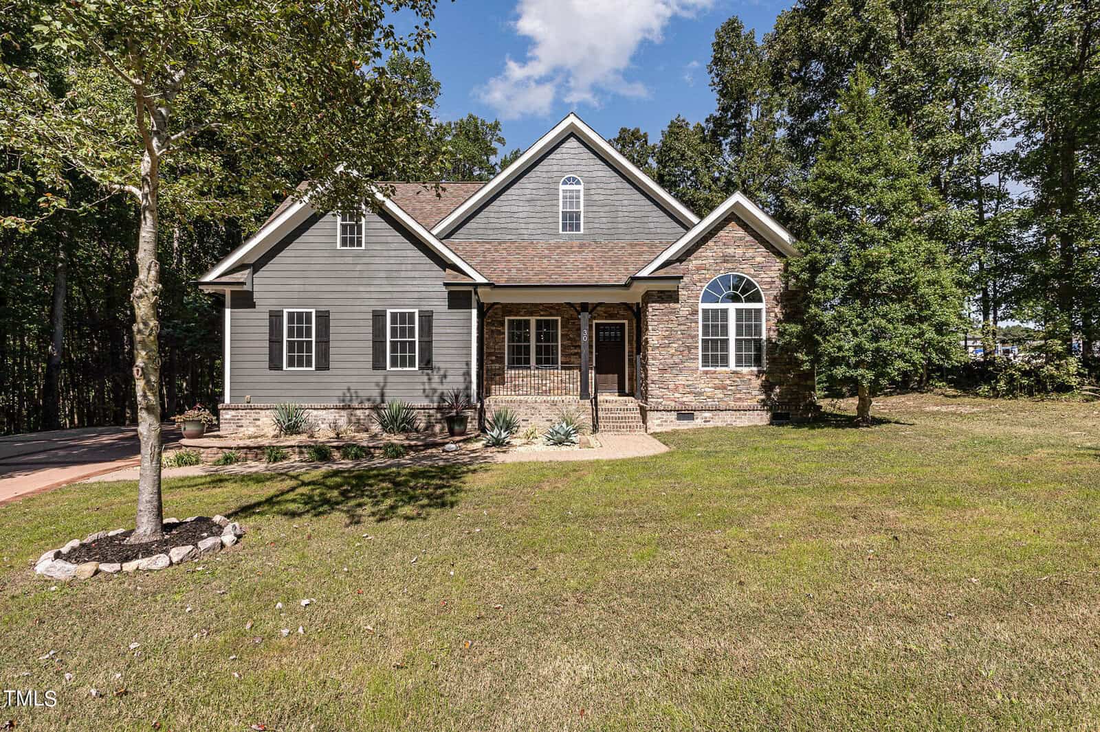 A gray and stone single-story house with a front porch, arched window, and gabled roof sits on a large grassy lawn with trees in the background under a blue sky with scattered clouds.