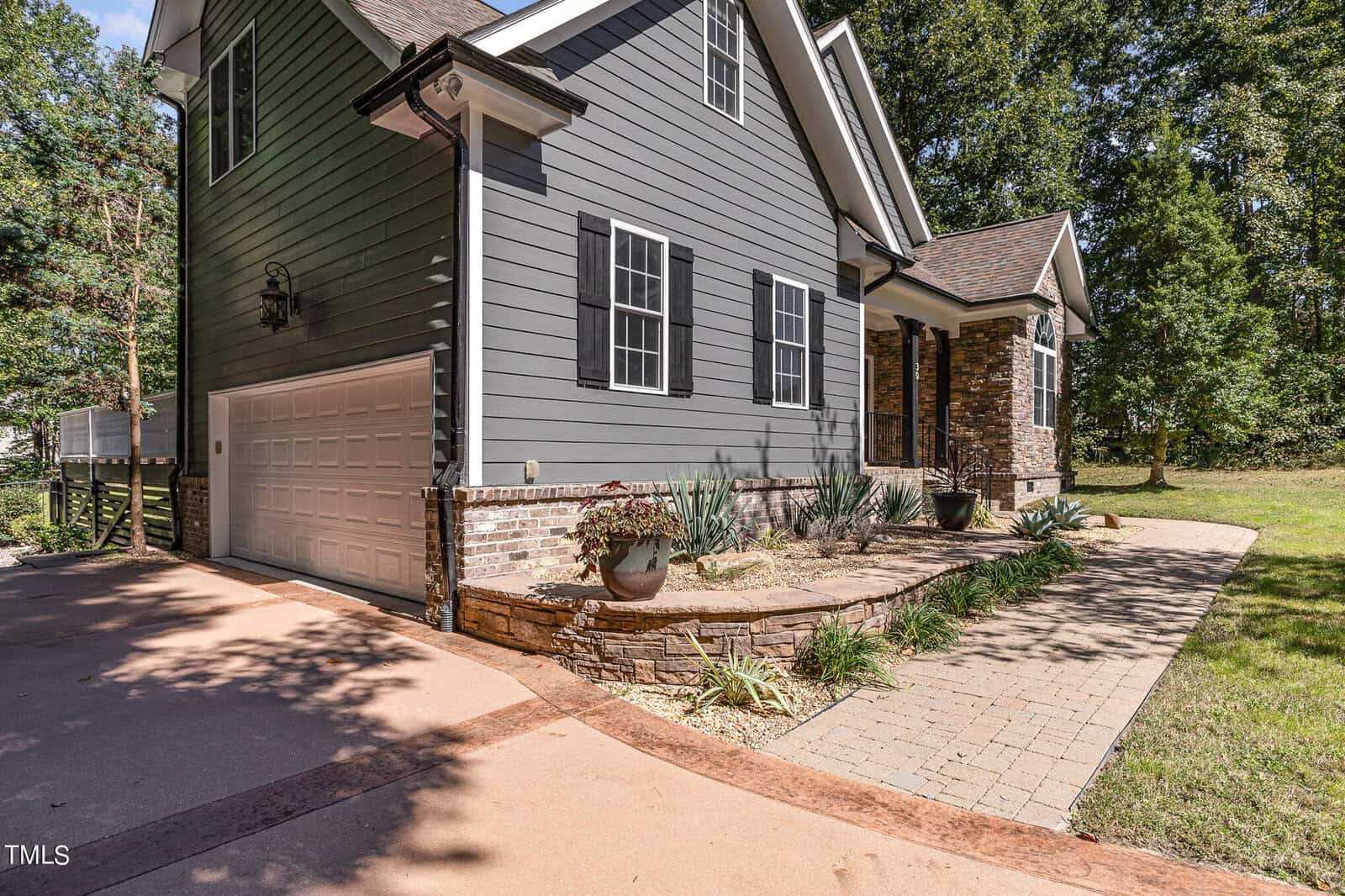 A modern house with gray siding, black shutters, and a two-car garage. The front yard features a paved walkway, stone landscaping, potted plants, and a neatly manicured lawn with surrounding trees.