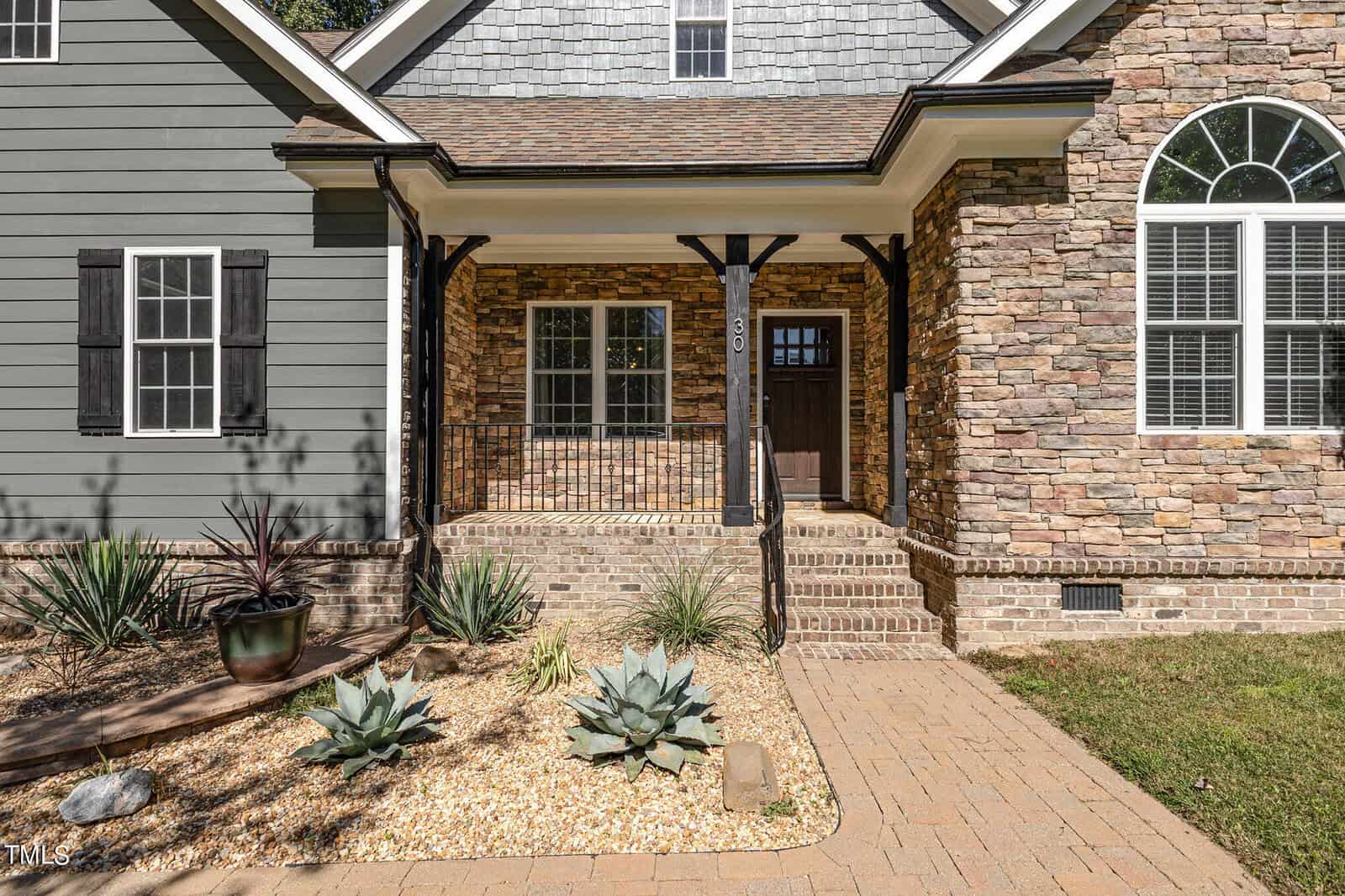 A house exterior with stone and gray siding, a brown front door, arched window, small porch with black railing, brick steps, and a landscaped front yard with succulents and gravel.