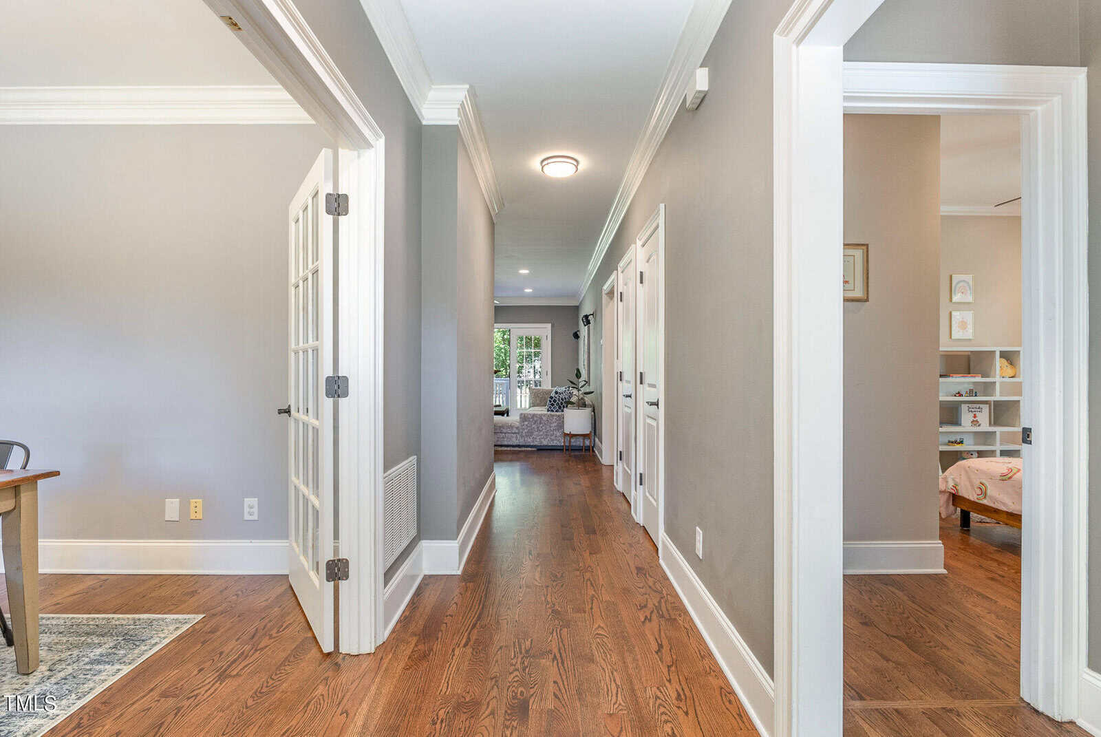A hallway with hardwood floors, gray walls, and white trim leads to a bright living area. On the left is a room with French doors and on the right, an open doorway reveals a child's bedroom with a bookshelf.