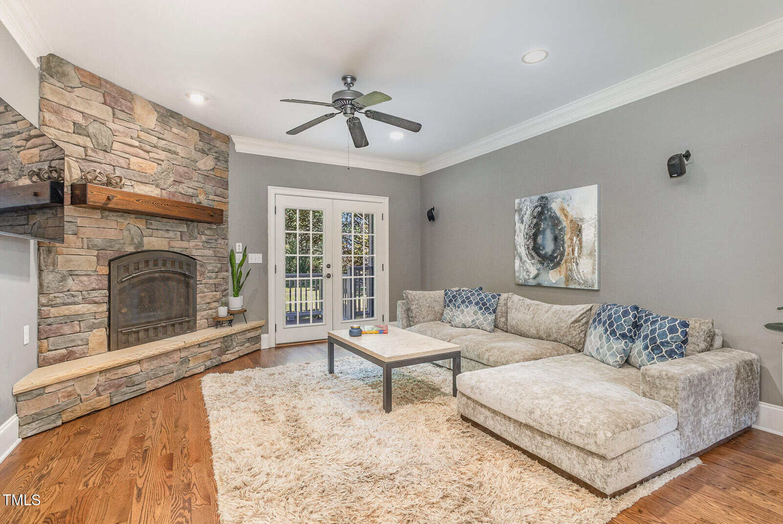 A cozy living room with a stone fireplace, wood mantel, beige sectional sofa with patterned pillows, a shaggy rug, and a coffee table; double glass doors open to the outside. Walls are gray with modern art and crown molding.