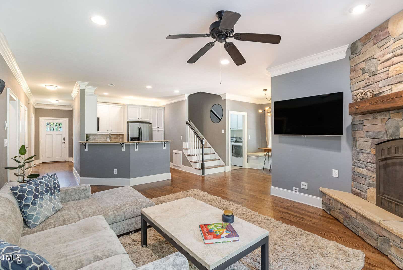 A cozy living room with a sectional sofa, a stone fireplace, wall-mounted TV, and a ceiling fan. The room opens to a kitchen with white cabinets, a breakfast bar, and hardwood floors throughout. Stairs lead to the upper level.