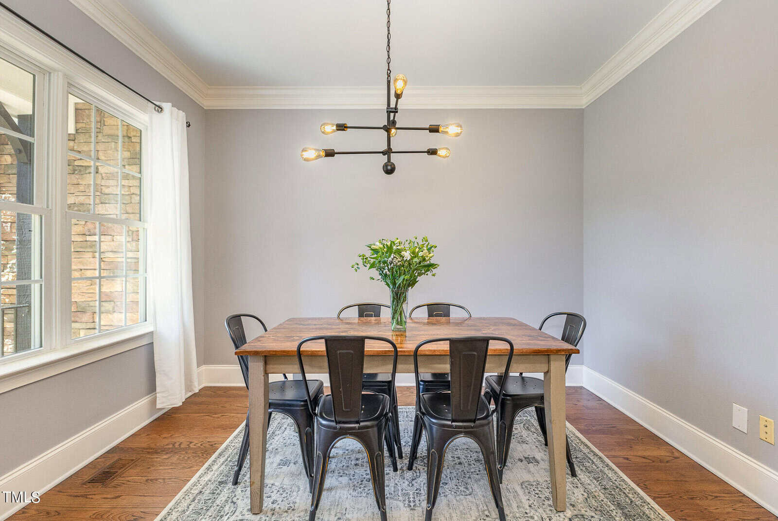 A dining room with a wooden table, six black chairs, a modern black chandelier, a vase of green flowers, hardwood floors, a gray area rug, gray walls, and a large window with white curtains.
