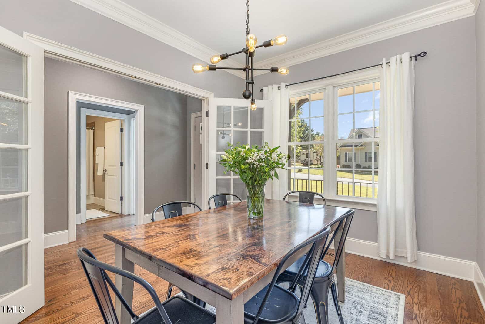 Bright dining room with a rustic wooden table, six black chairs, a modern chandelier, and large windows with white curtains letting in natural light. A vase with green leaves sits at the center of the table.