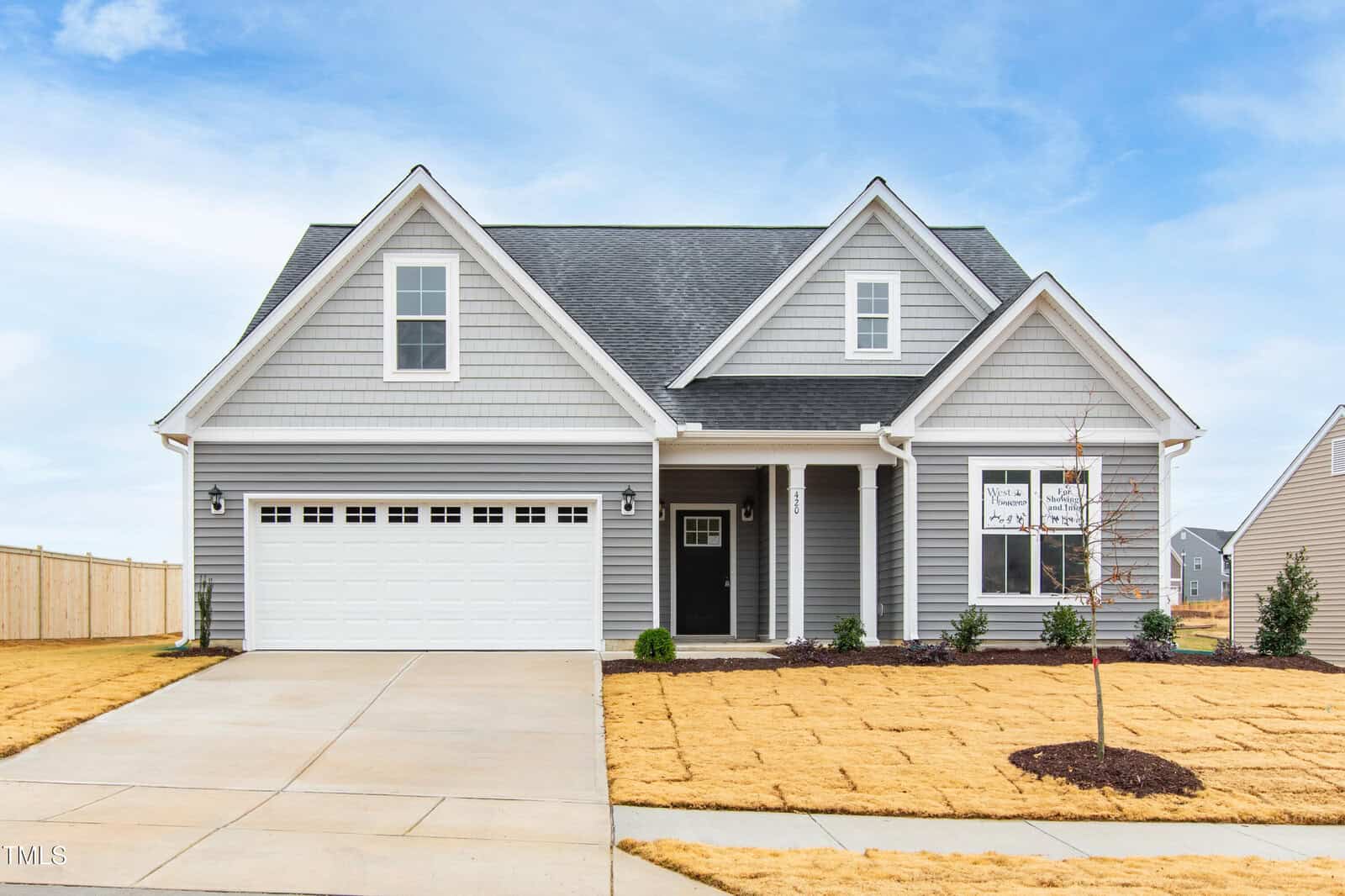 A newly constructed, two-story gray house with white trim, a double garage, and a small front porch. The yard is freshly seeded with straw covering the grass, and there is a young tree in the front lawn.