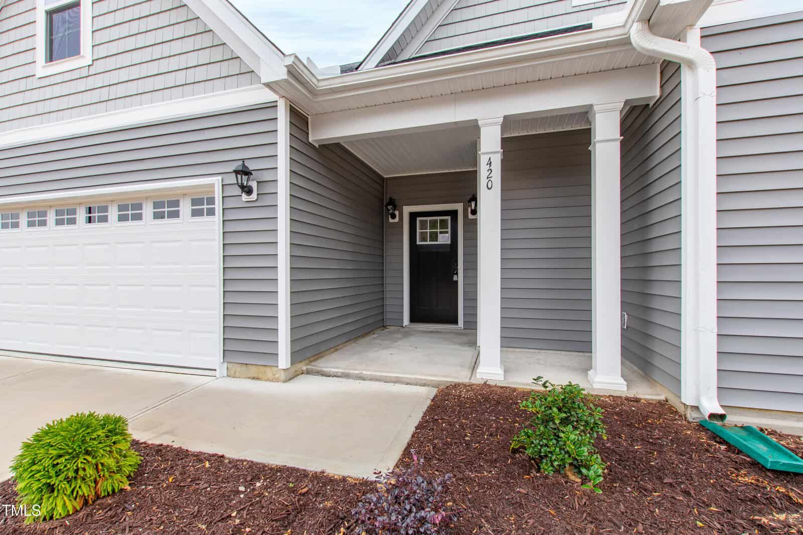 A covered front entryway of a house with gray siding, white trim, a black front door, two exterior lights, a column, a garage door on the left, and small landscaped bushes along a concrete path.