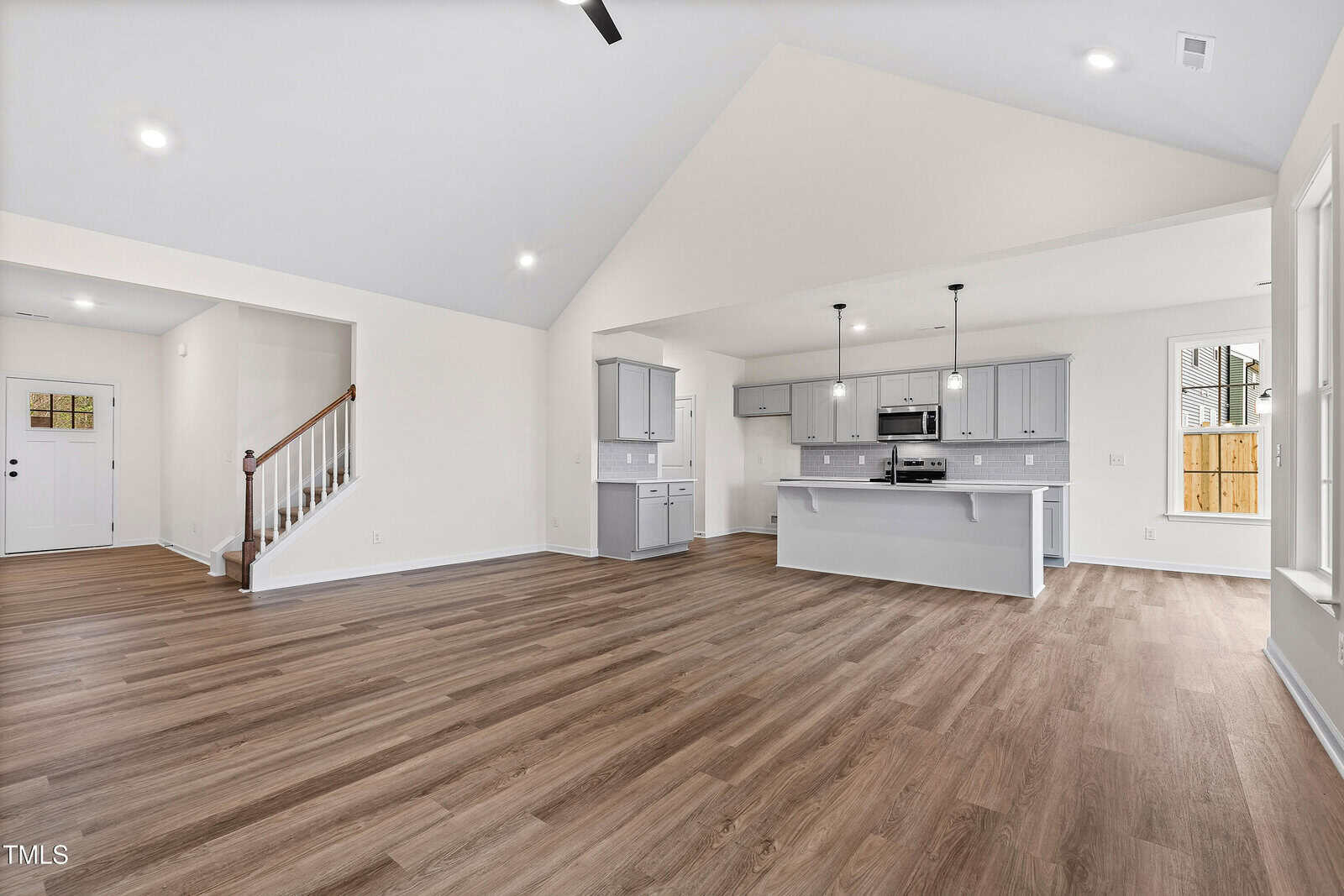 Open-concept living area with light wood flooring, vaulted ceiling, and recessed lighting. The kitchen features gray cabinets, an island, stainless steel appliances, and pendant lights. Stairs lead to an upper level.