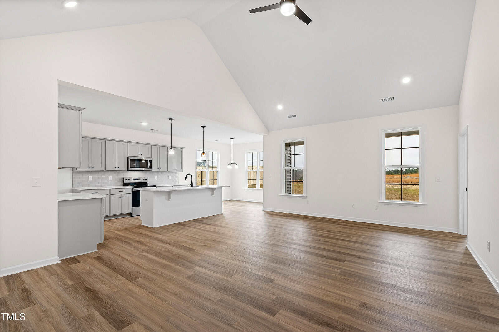 A spacious, empty open-concept living area with light wood flooring, white walls, large windows, and a modern kitchen featuring gray cabinets, stainless steel appliances, and a central island with pendant lighting.