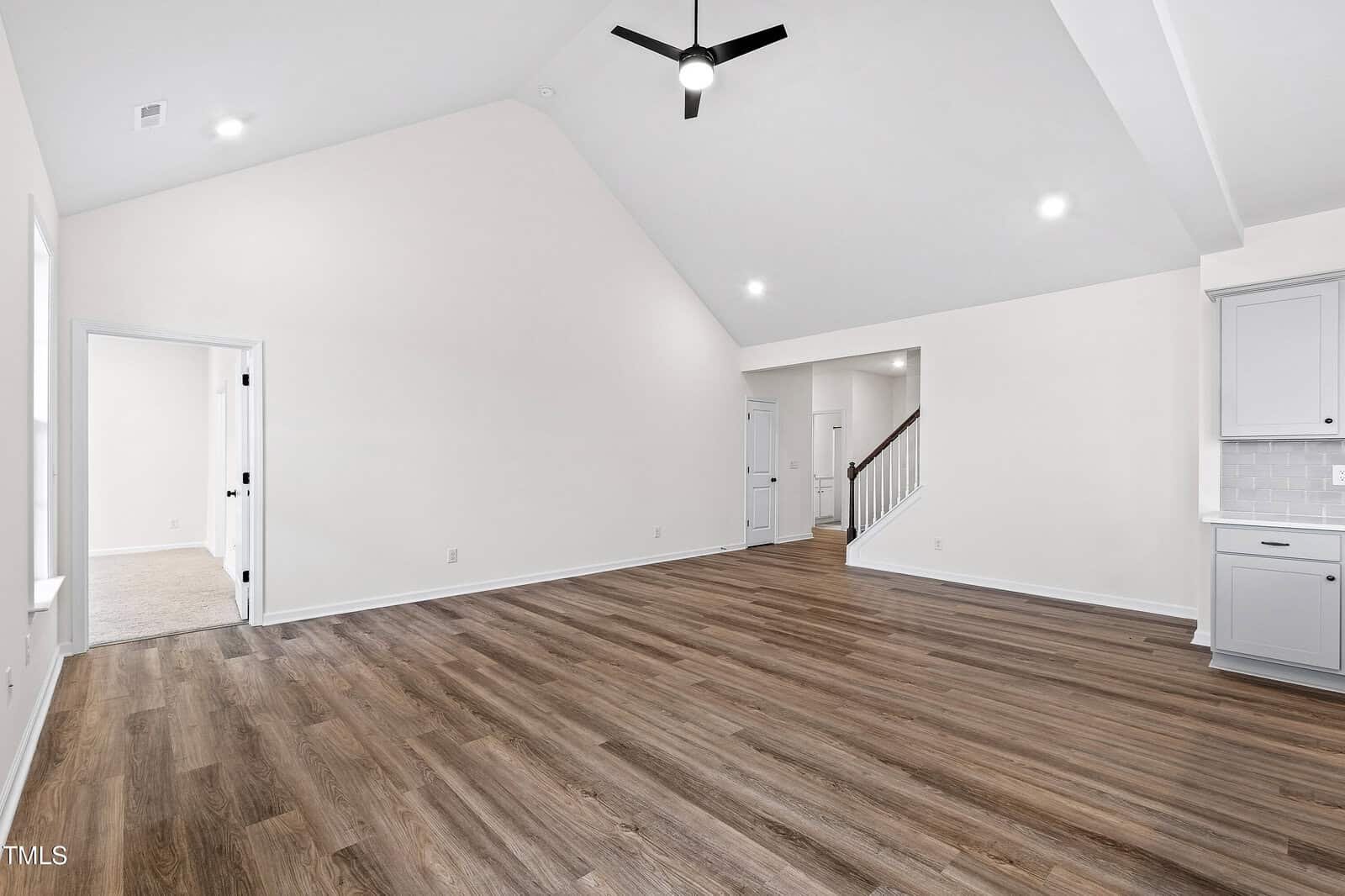 Spacious, empty living room with high vaulted ceiling, wood-style flooring, neutral walls, recessed lighting, and a ceiling fan. Visible doorways lead to adjoining rooms and a staircase. Part of a kitchen is visible on the right.