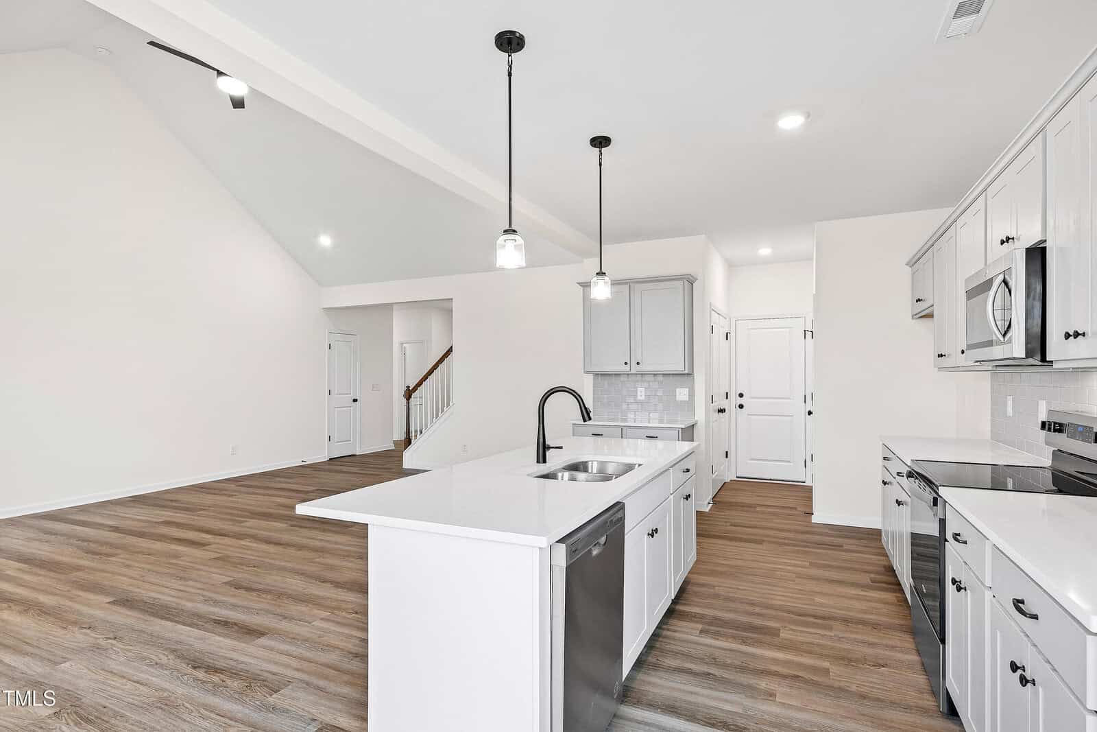 A modern kitchen with white cabinets, stainless steel appliances, a central island with a sink, and pendant lighting. The space opens to a living area with wood flooring and high ceilings.