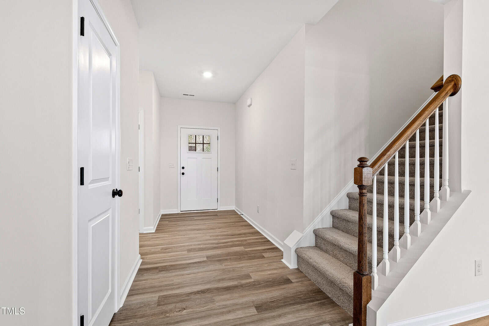 A hallway with light wood flooring, white walls, a closed white door on the left, a staircase with carpeted steps and a wooden handrail on the right, and a white front door with a small window at the far end.