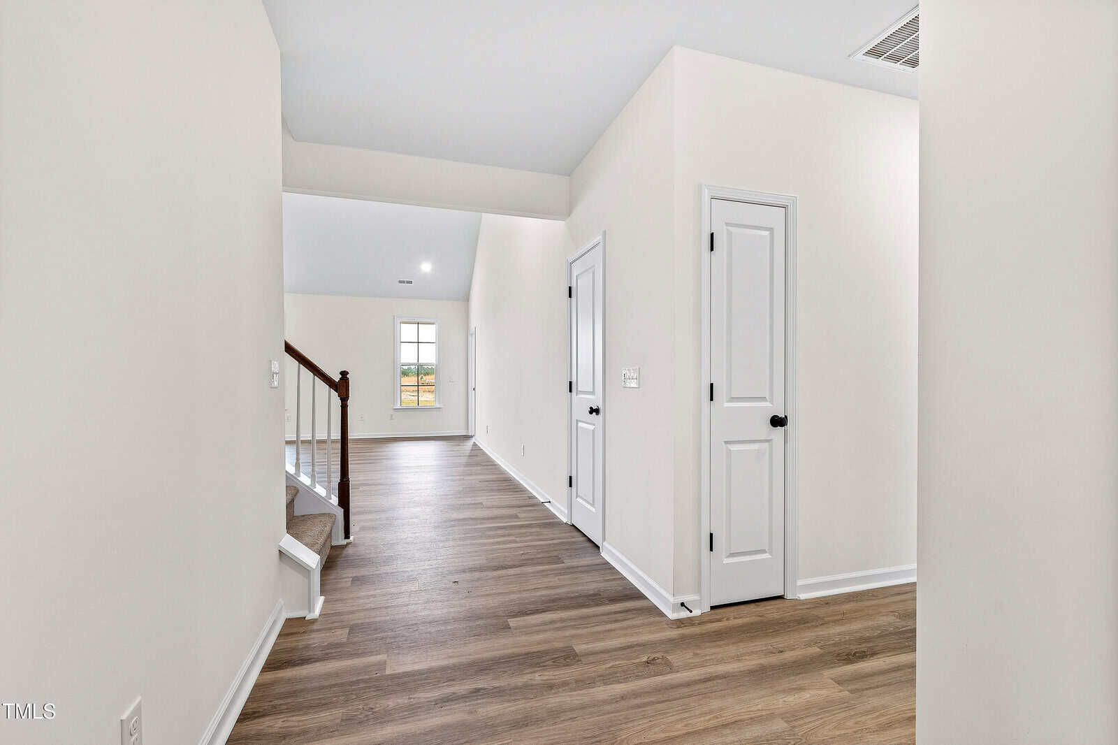 A hallway in a modern home with light-colored walls and wood-style flooring. There are white doors with black handles, a staircase with a wooden handrail, and a window at the end of the hall.