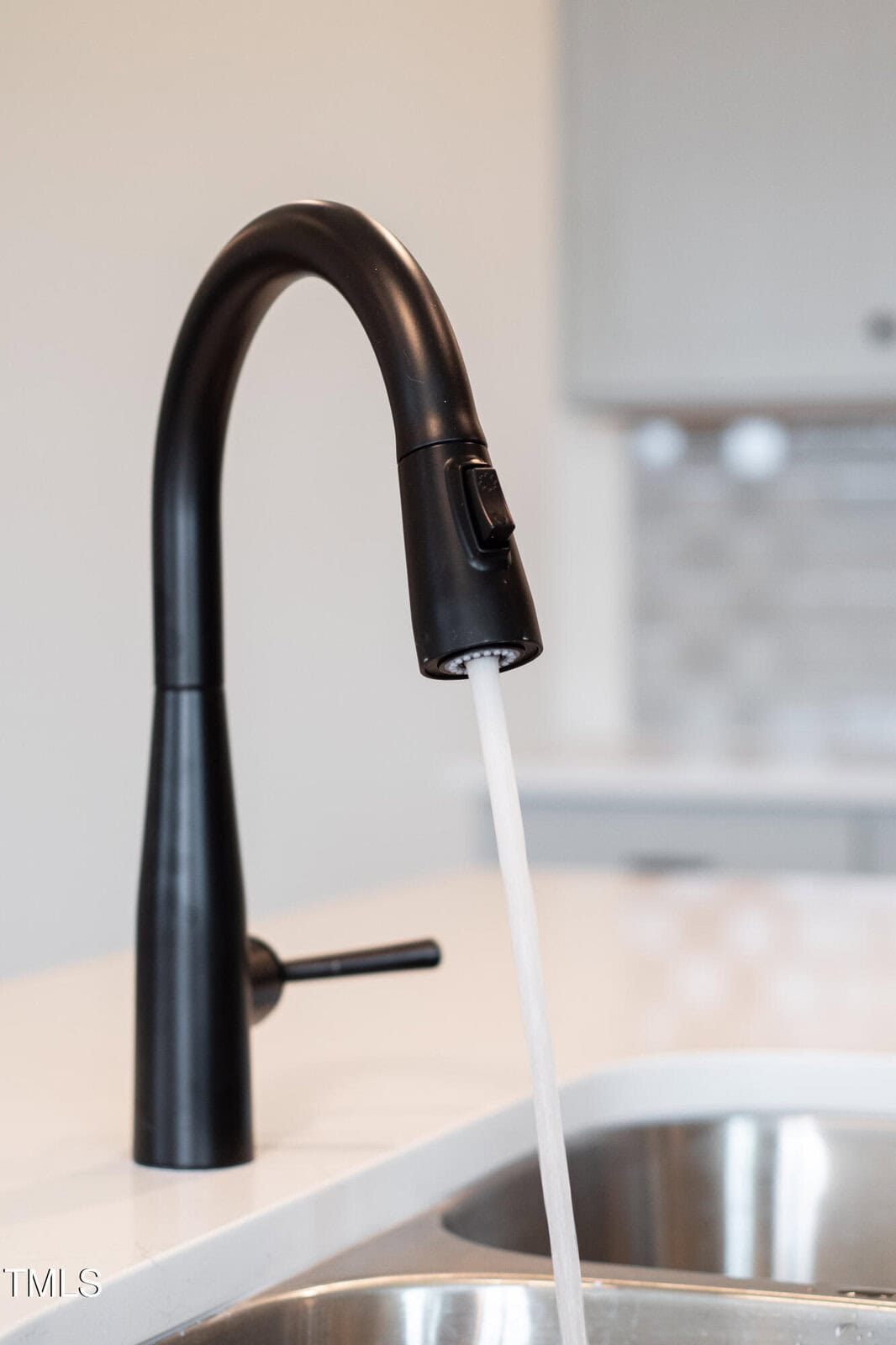 A black kitchen faucet with a curved neck and single lever handle has water running into a stainless steel sink on a white countertop; a blurred backsplash is visible in the background.