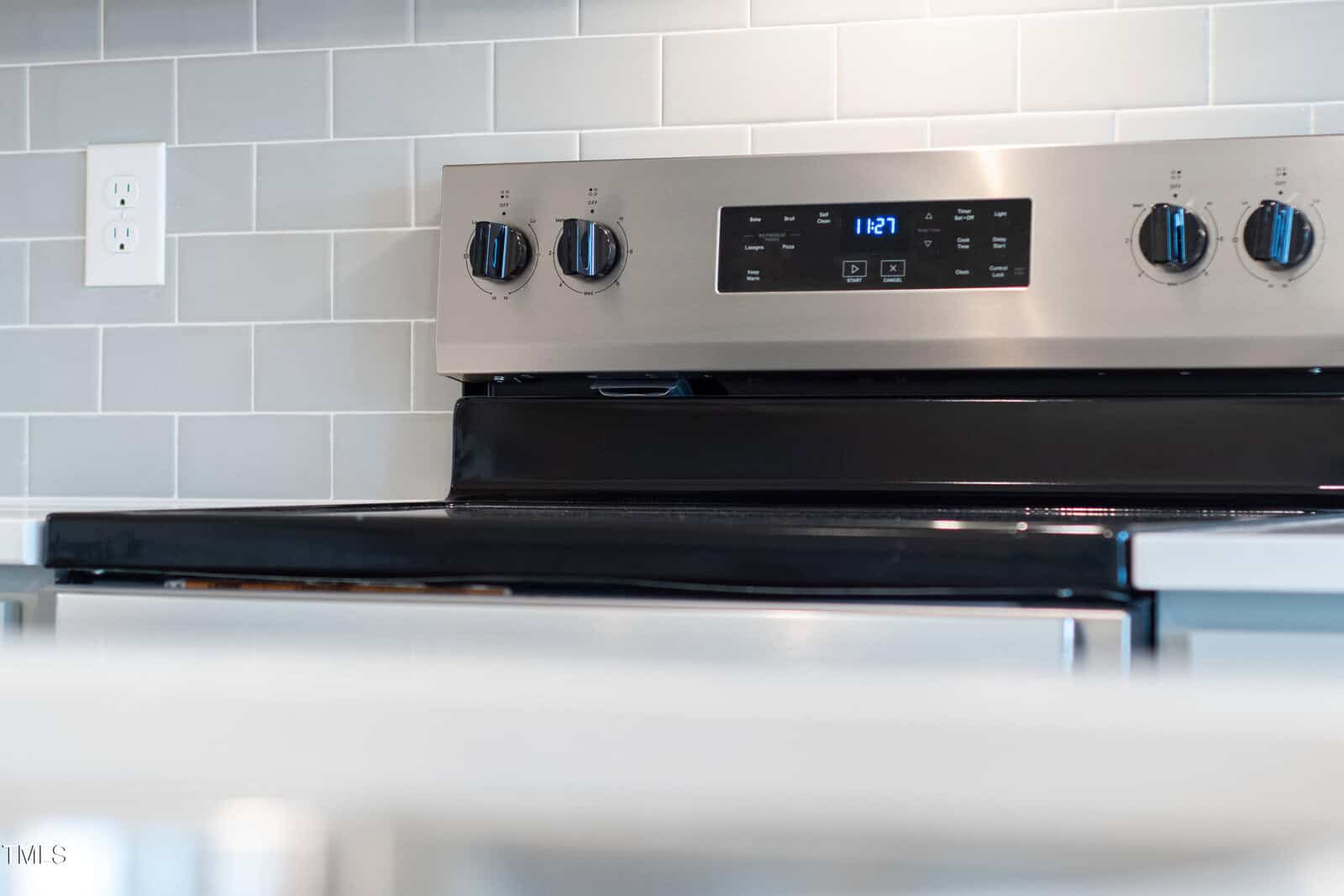 A close-up of a stainless steel electric stove with black knobs and digital clock, set against a light gray subway tile backsplash.