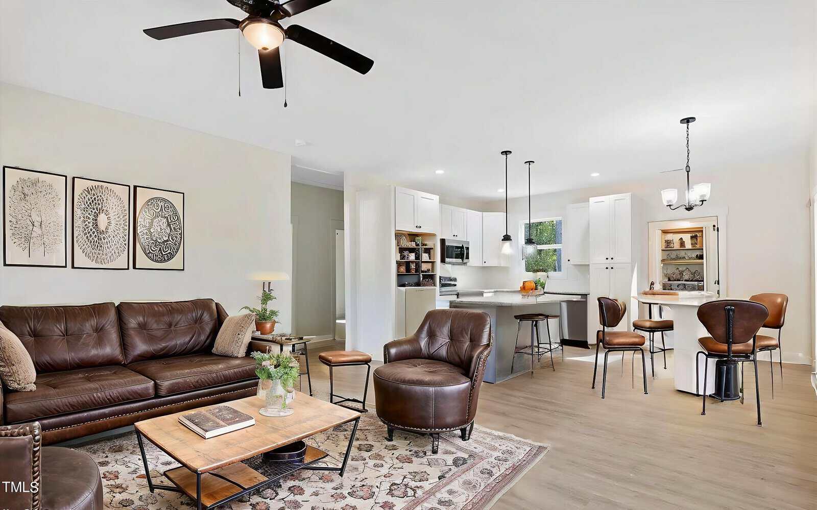 A modern open-plan living room and kitchen with brown leather furniture, a patterned rug, white cabinetry, pendant and ceiling lights, and bar stools at a kitchen island. Neutral colors and decorative wall art create a cozy atmosphere.