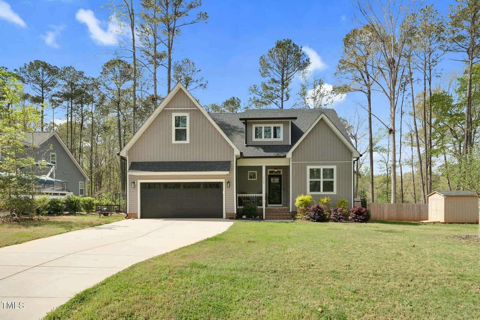 A modern two-story house with gray siding, a black roof, and a double garage, set on a spacious lawn with a driveway. Trees and a small shed are visible in the background under a clear blue sky.