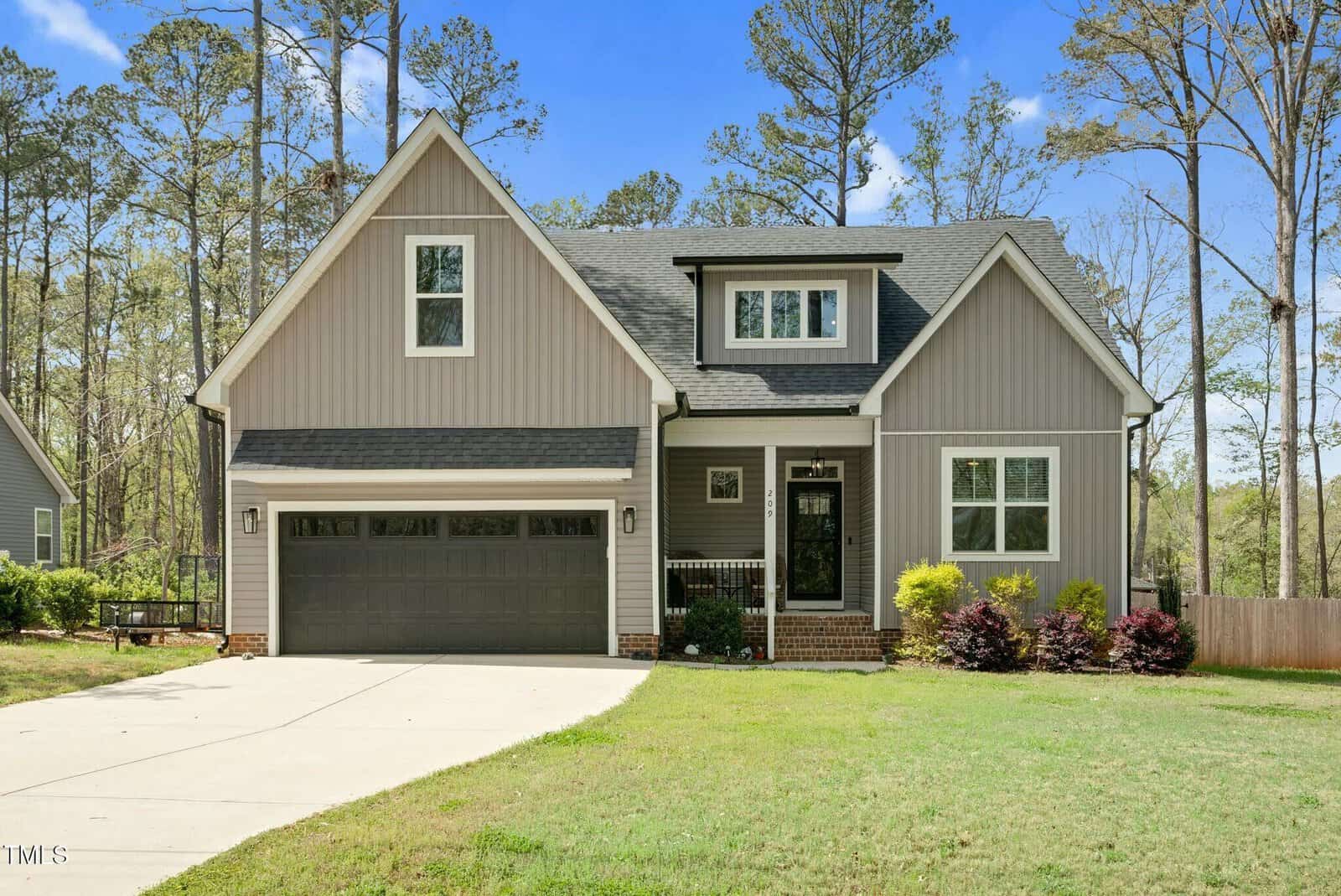 A two-story gray house with white trim, a dark gray double garage, and a small front porch with black railing. The house has a large driveway and a well-kept lawn, surrounded by tall trees and landscaping.