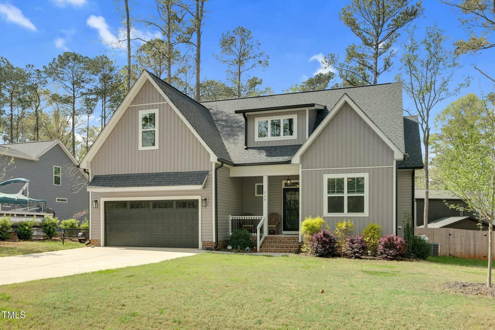 A two-story gray house with white trim, a dark gray double garage door, a small front porch, and shrubs in the front yard, set against a backdrop of tall trees and a clear blue sky.