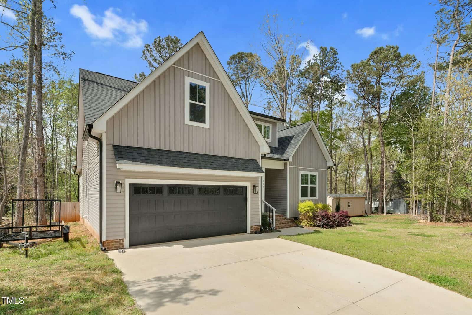 A modern two-story house with gray siding, a dark gray garage door, and a steep gable roof, set among tall trees with a wide concrete driveway in front and a small trampoline on the left.