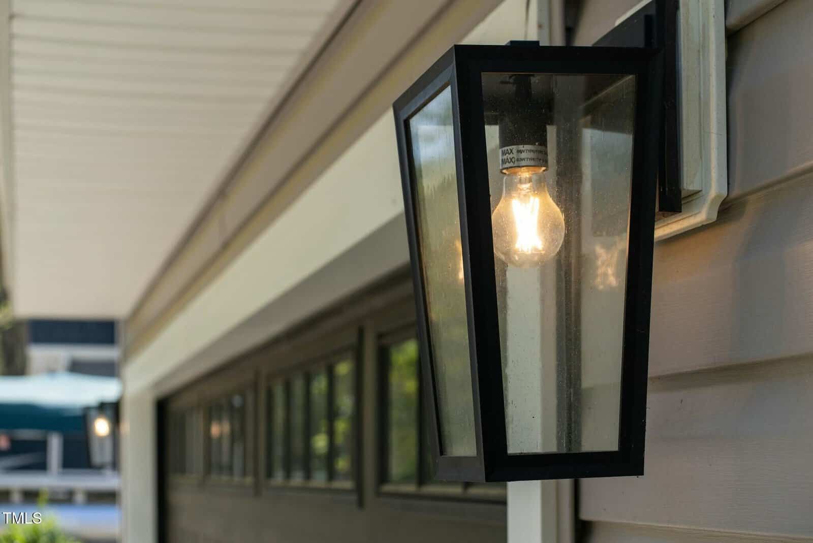 A lit outdoor wall-mounted lantern with a black frame is attached to the siding of a house, positioned near a dark garage door.