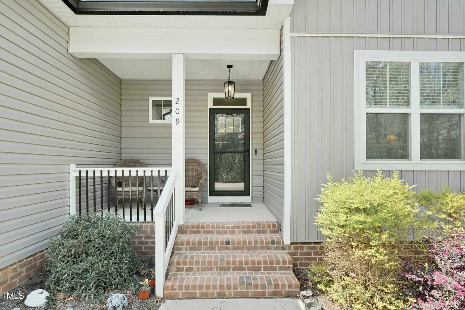 A brick front porch with a black door, white trim, house number 209 on a post, a hanging lantern light, two wicker chairs to the left, gray siding, and a shrub in the landscaped area.