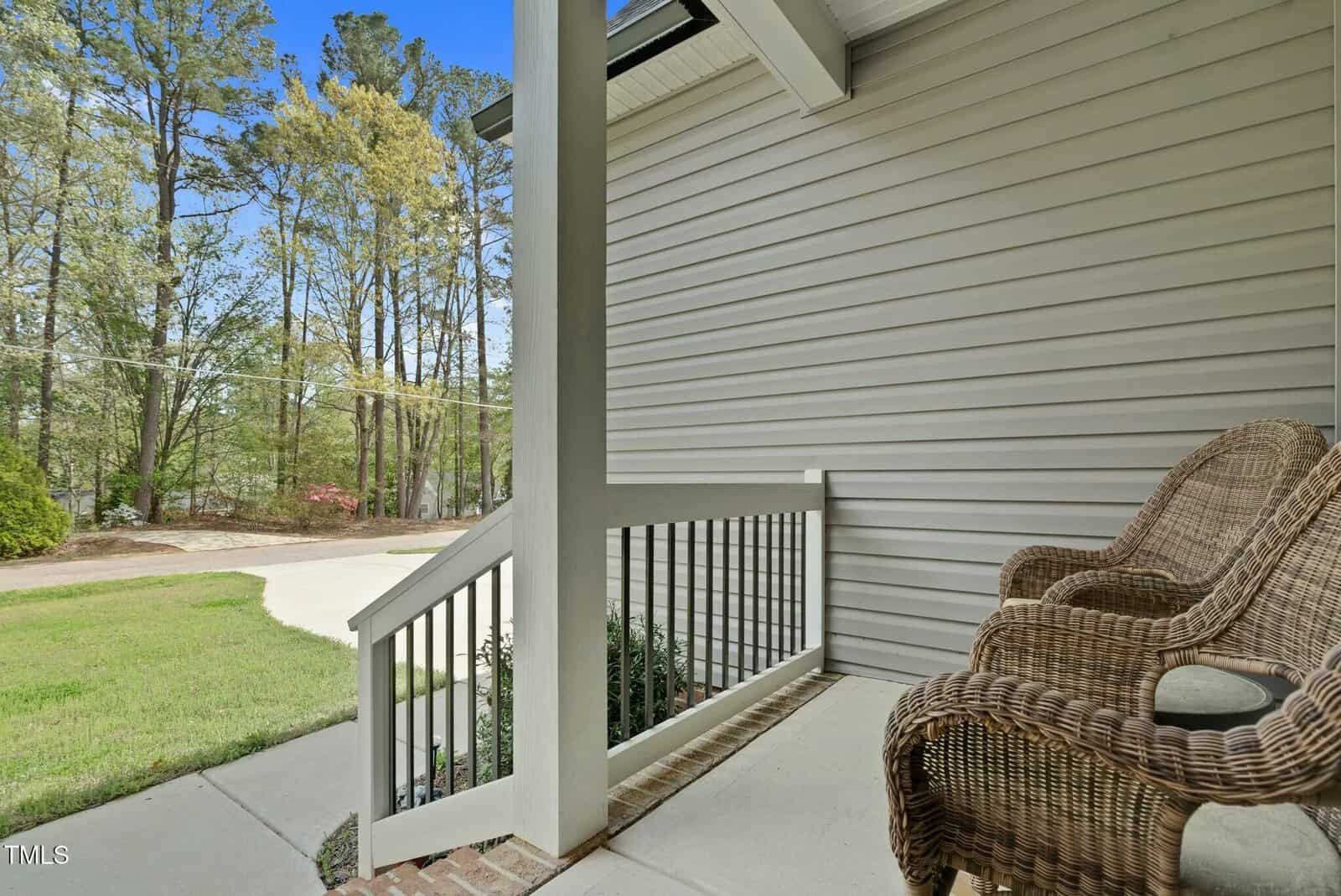 Two wicker chairs sit on a covered porch with beige siding and a white railing. The porch overlooks a green lawn, driveway, and trees in the background under a clear sky.