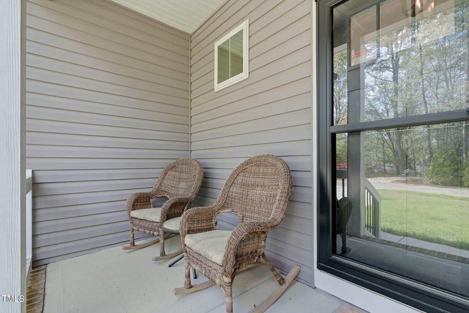 Two wicker rocking chairs with cushions are placed side by side on a covered porch with beige siding. A small rectangular window is on the wall, and a glass door is partially open, showing the porch steps and yard outside.