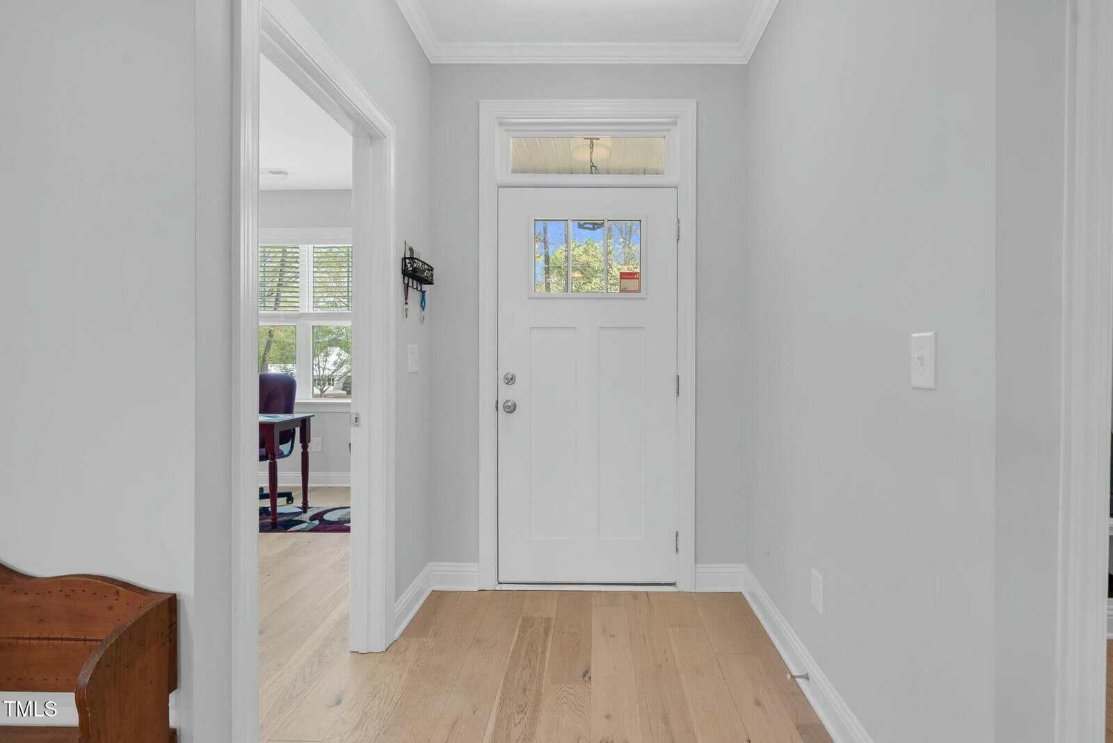 A hallway with light gray walls, light wood flooring, and a white front door with glass panes. To the left, an open doorway leads to a room with a desk and large windows.