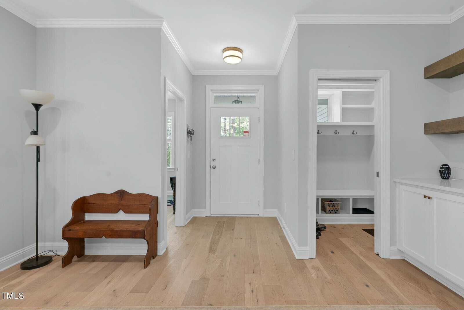Entryway with light wood floors, white walls, a wooden bench, a floor lamp, built-in shelves, baskets, and a white front door with glass panels allowing natural light inside.