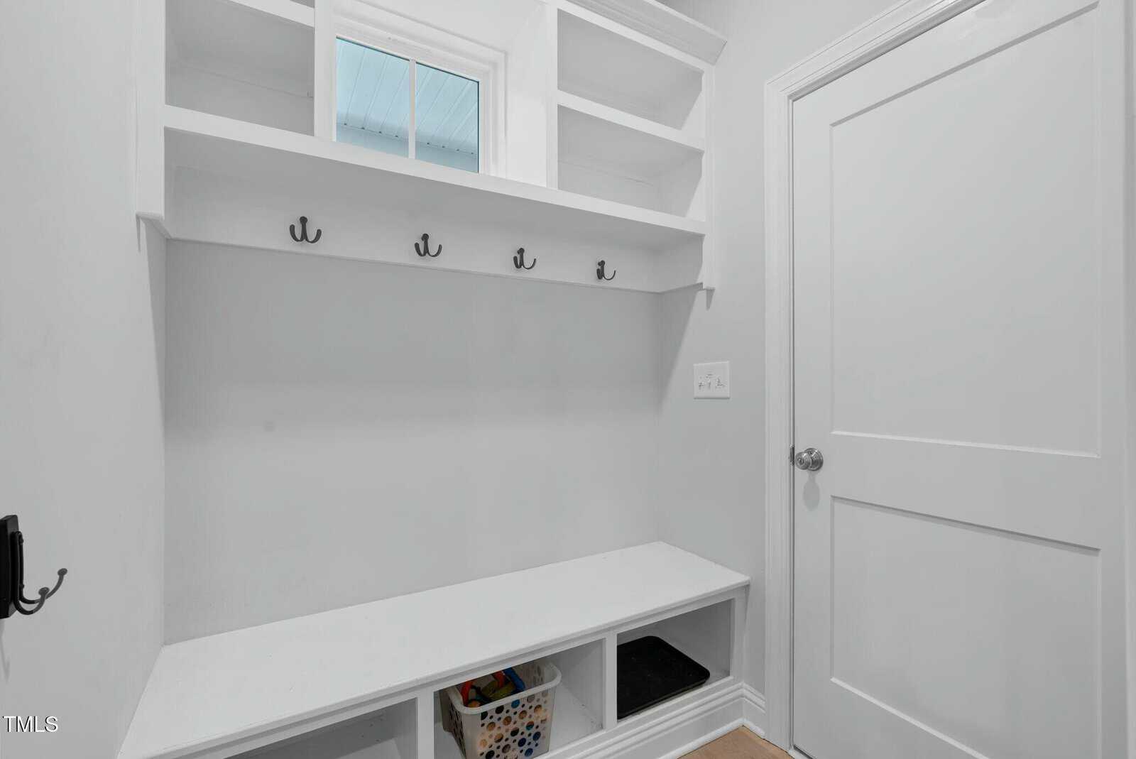 White mudroom with open shelves, coat hooks, and a bench. Storage cubbies below the bench hold a basket with colorful items. A closed white door is on the right, and a small window is above the shelves.