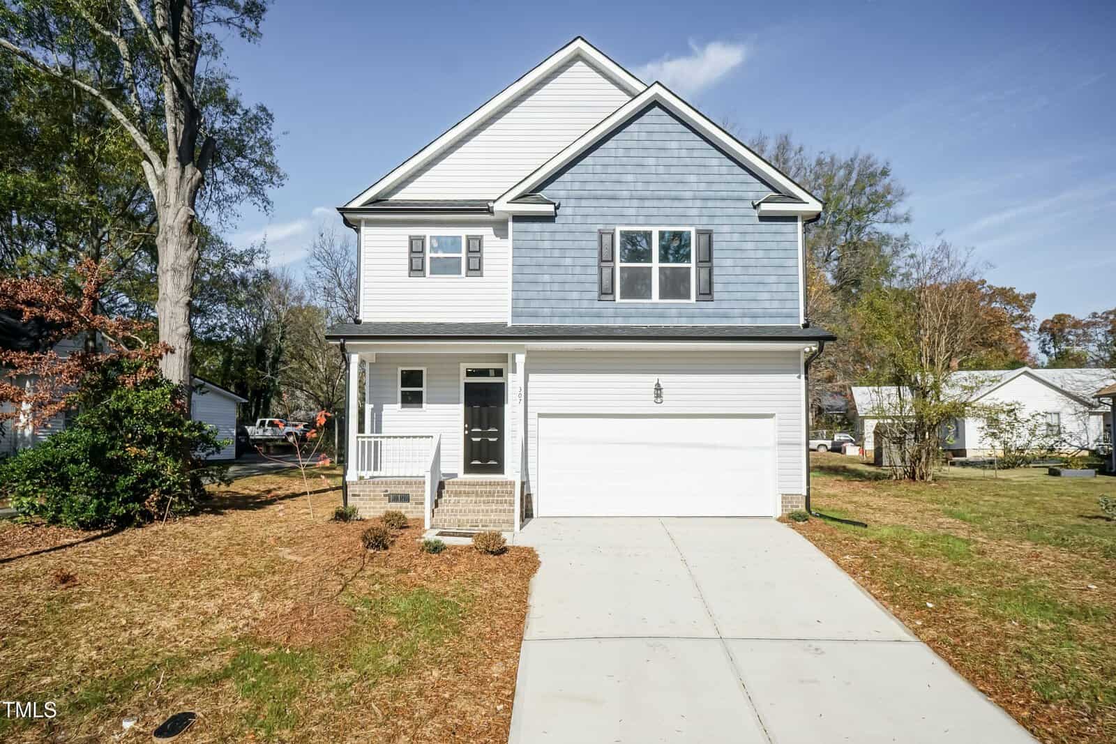 A two-story suburban house with a blue and white exterior, a covered front porch, black front door, attached two-car garage, and a concrete driveway, surrounded by grass and trees under a clear sky.