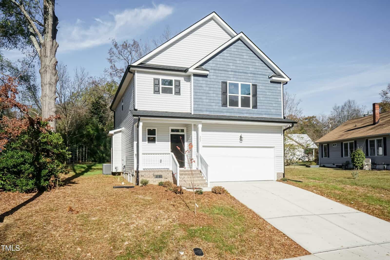 A two-story house with gray siding and blue accents, attached garage, white front door, small front porch, and a concrete driveway. There is minimal landscaping with patchy grass and a few shrubs.