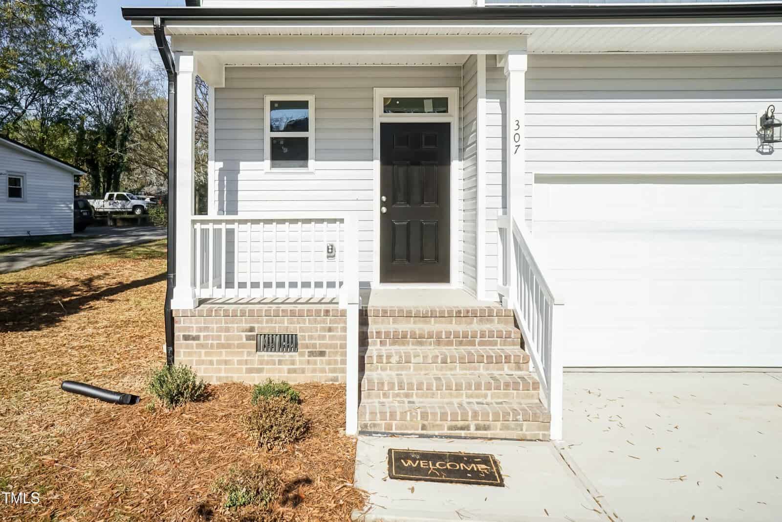 A small brick porch with white railings leads to a black front door on a light gray house. The house number 307 is visible next to the door, and a welcome mat is on the porch. Shrubs and mulch are in front.