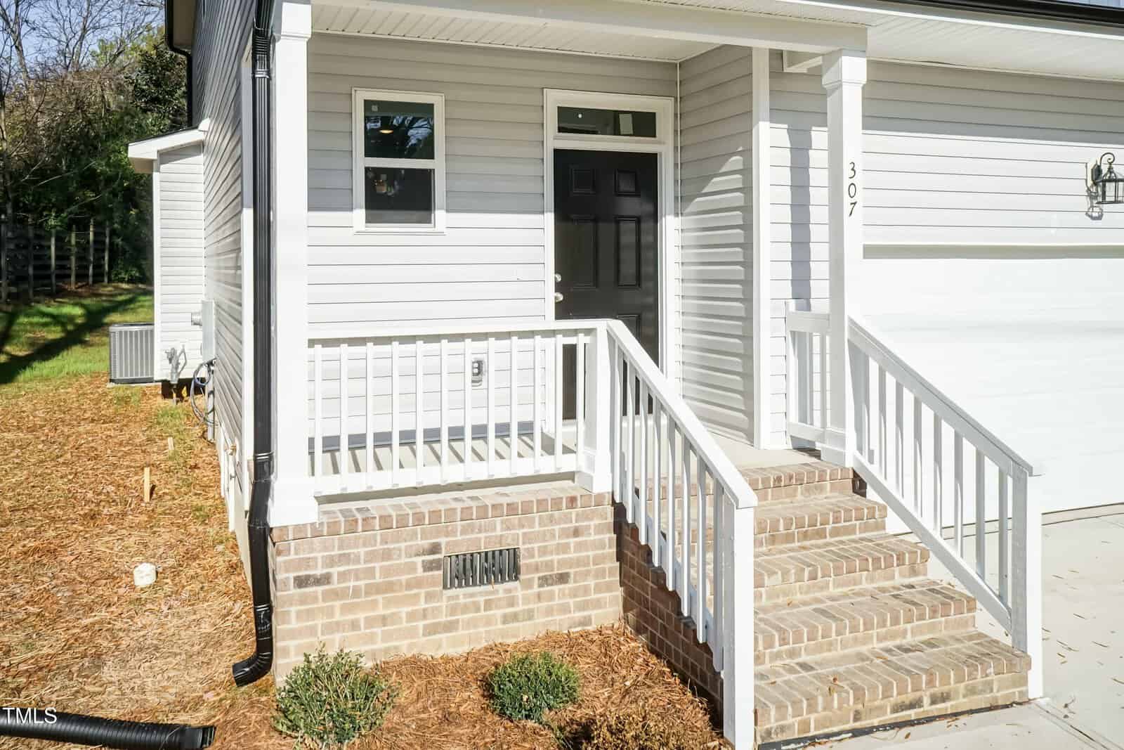 A small front porch with white railings and steps leads to a black door on a light gray house with brick foundation. A garage door is next to the porch, and there are some shrubs planted in front.