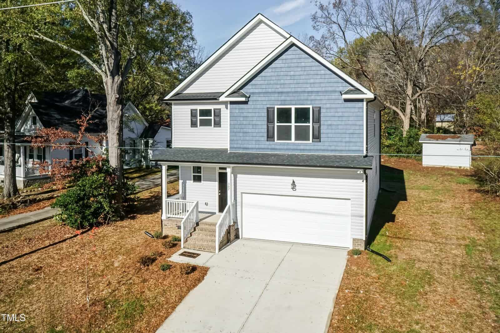 A two-story house with blue and white siding, a front porch, and an attached two-car garage. The property has a concrete driveway, sparse grass, and trees in the yard, with a small shed visible in the backyard.