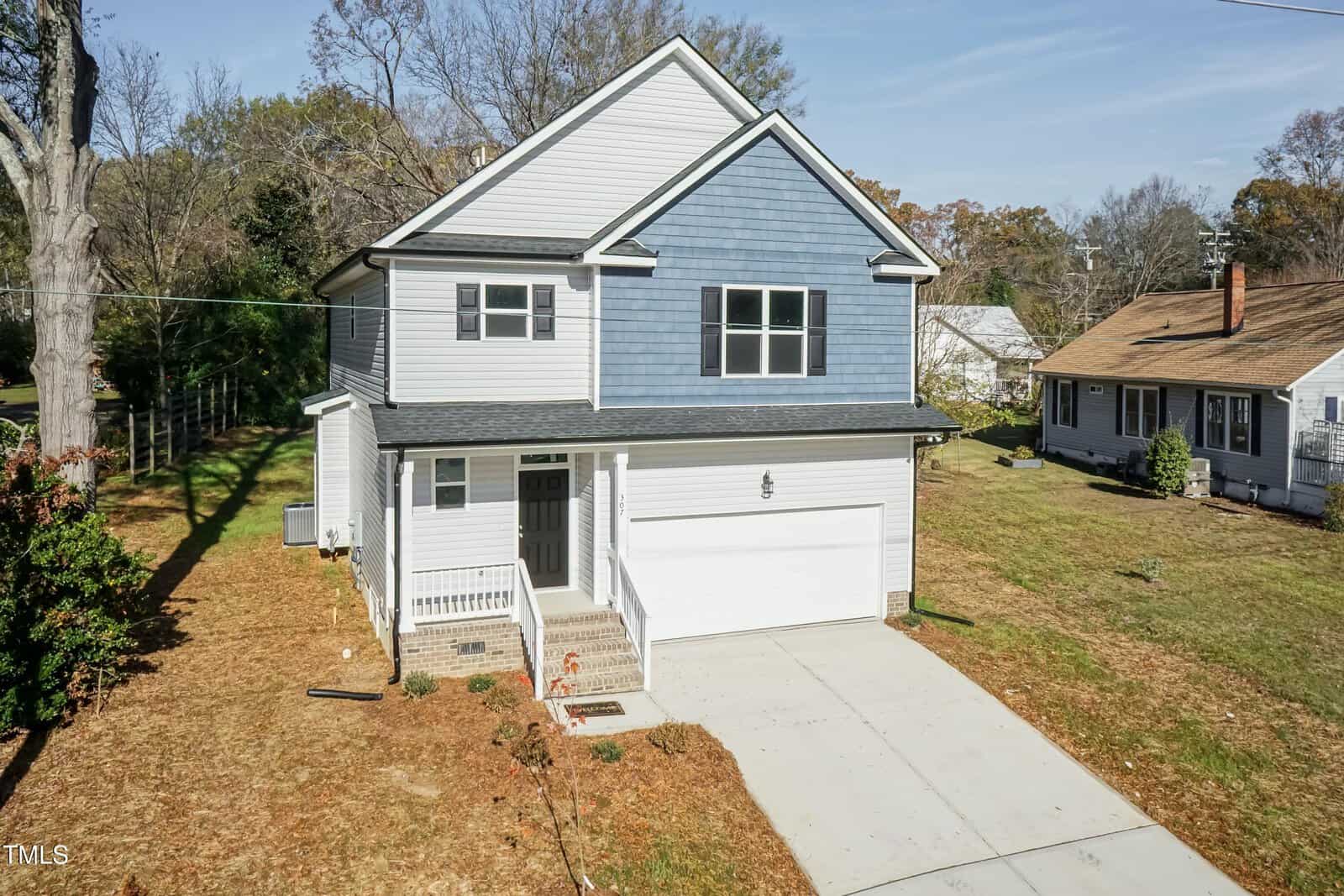 A two-story house with blue and white siding, a front porch, and a white garage door. The home features a concrete driveway and steps leading to the entrance, surrounded by a grassy yard and neighboring houses.
