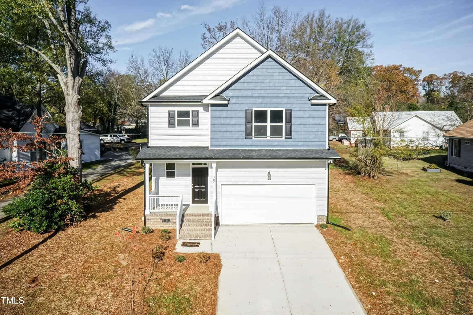 A two-story house with blue and white siding, an attached two-car garage, a small front porch, and a concrete driveway, surrounded by dry grass and a few trees in a suburban neighborhood.