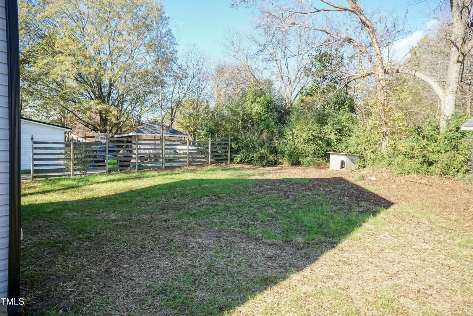 A backyard with a mix of green grass and bare patches, bordered by a wooden fence and trees. There is a small dog house in the back right corner, and neighboring buildings are partially visible.