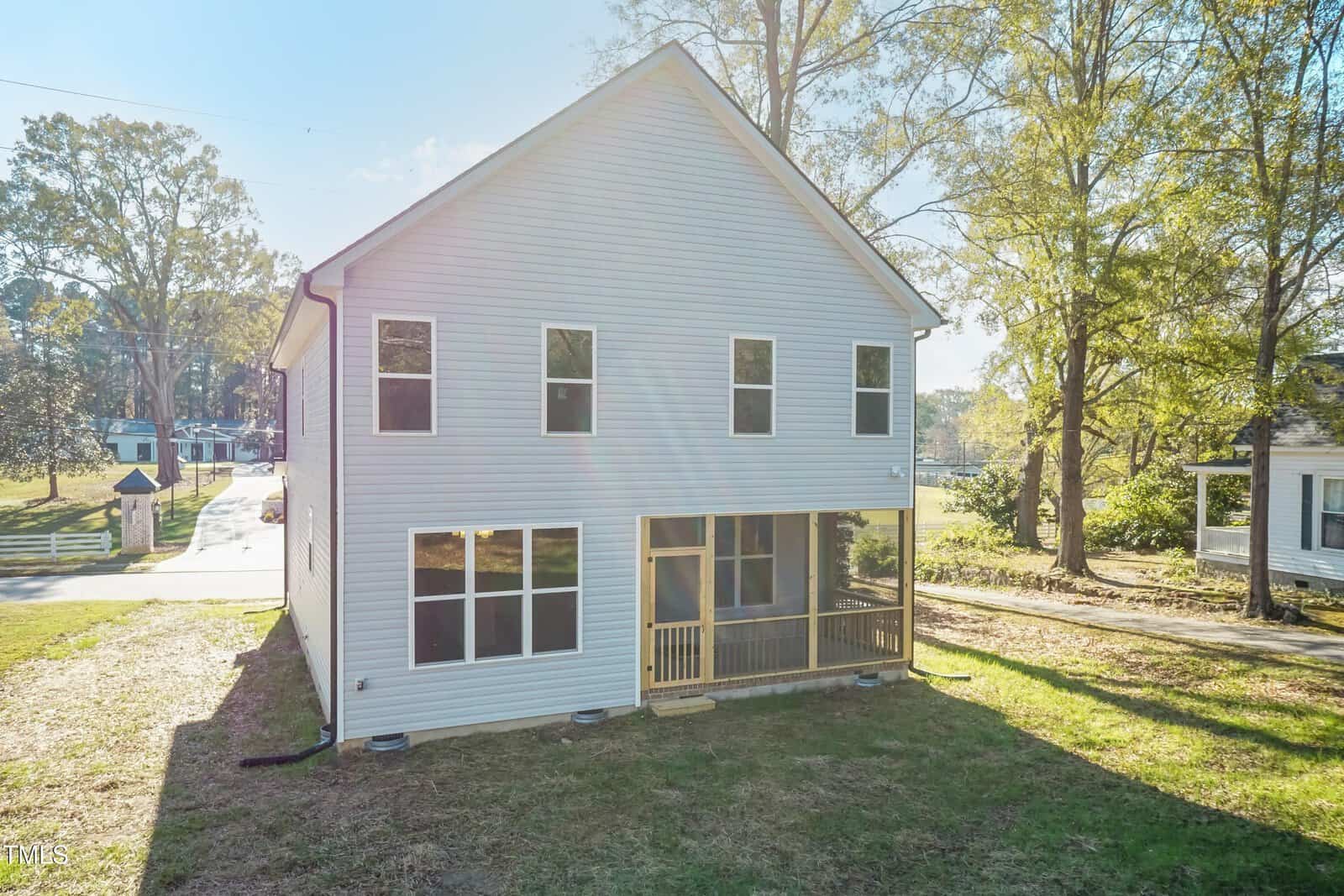 Two-story white house with multiple windows, a small screened porch, and a grassy yard, located on a corner lot with trees and a paved road nearby on a sunny day.