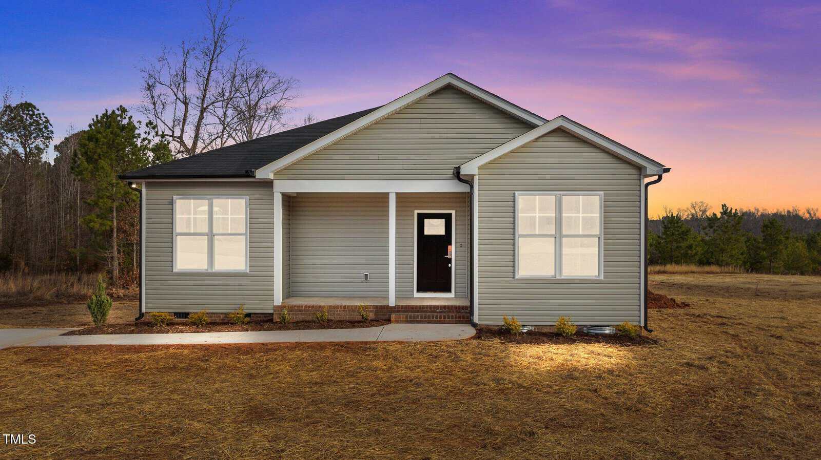 Single-story house with gray siding, white trim, and a small covered front porch. The yard is bare with some small shrubs, surrounded by trees. The sky is purple and orange, indicating sunset or sunrise.